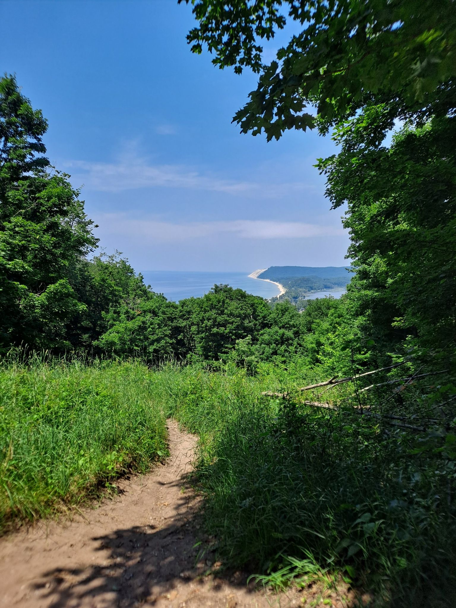 Sunny dirt path through green forest overlooking a large body of water. - Empire Bluff Trail