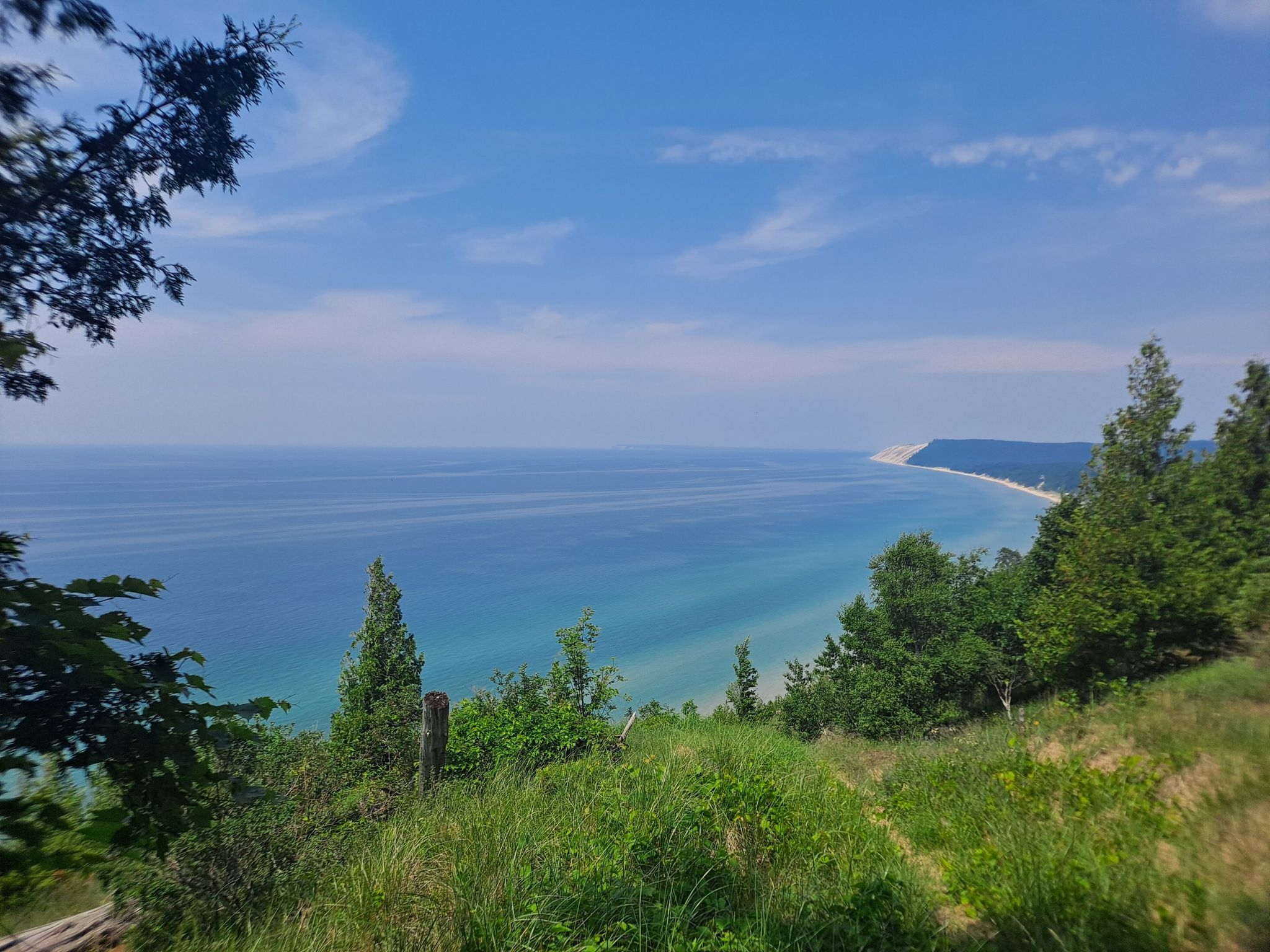Panoramic view of calm blue water and distant sandy dunes from a green hillside. - Empire Bluff Trail