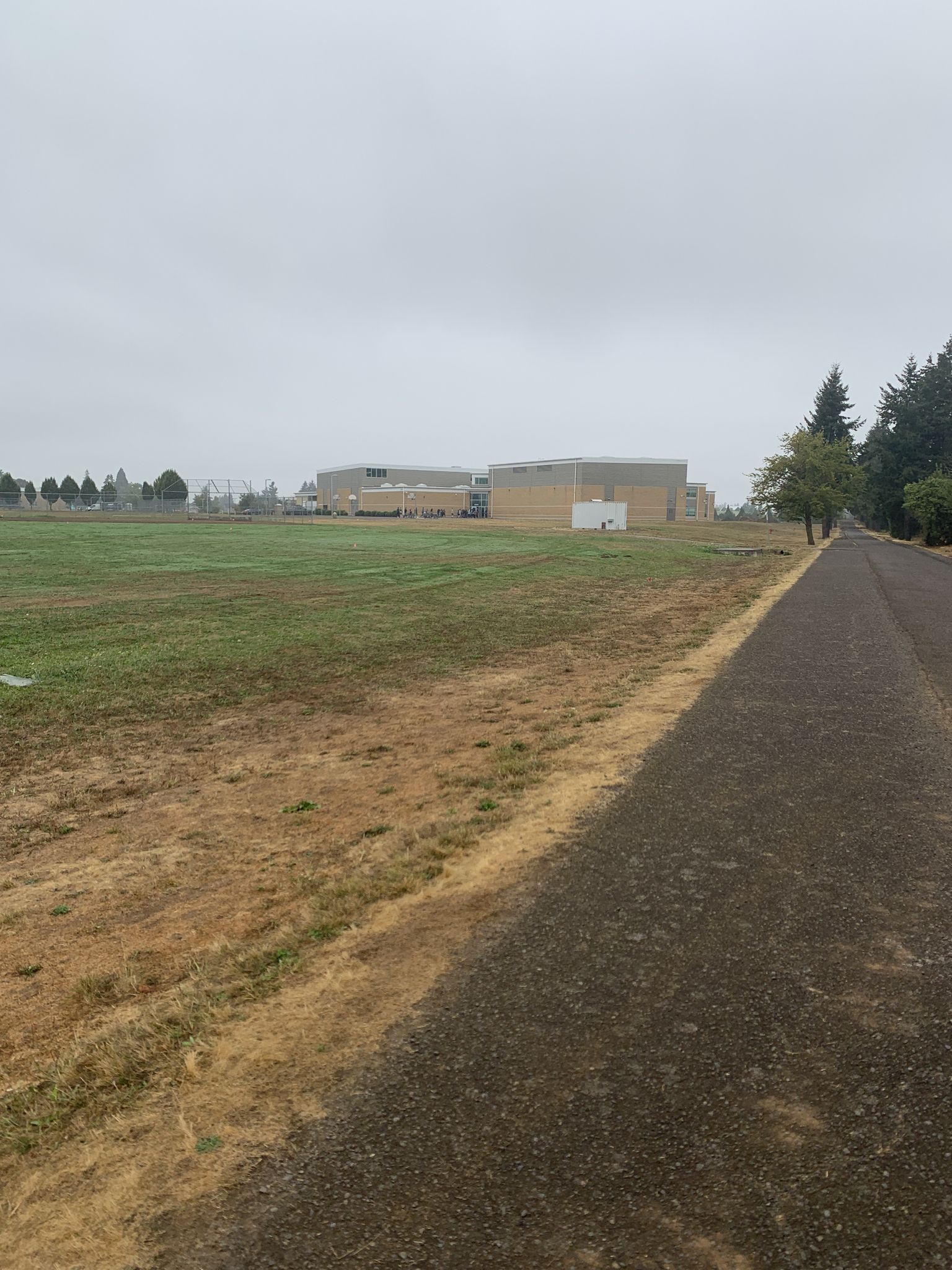 Paved path next to a grassy field with buildings under an overcast sky. - Logging County Road loop from Canby