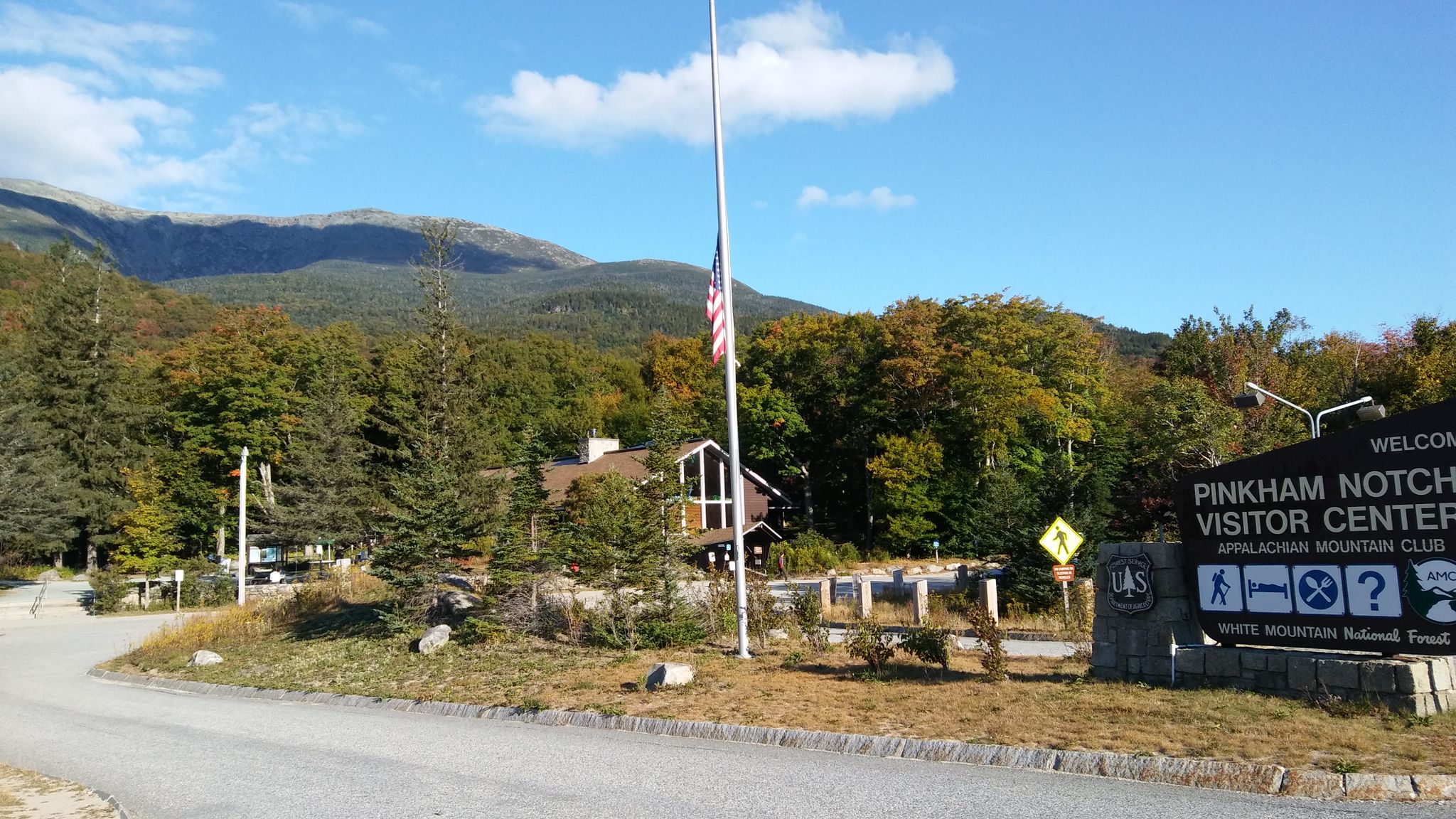 Roadside sign and flagpole in front of a building and forested mountains. - Gorham, Carroll, & Jackson loop