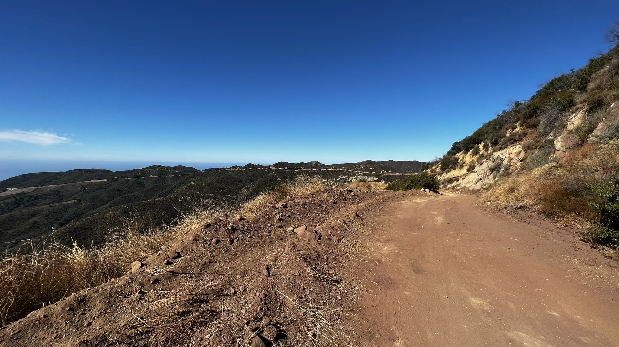 A dirt road on a hillside overlooking distant mountains and ocean under a clear sky. - Corral Canyon to Backbone Trail