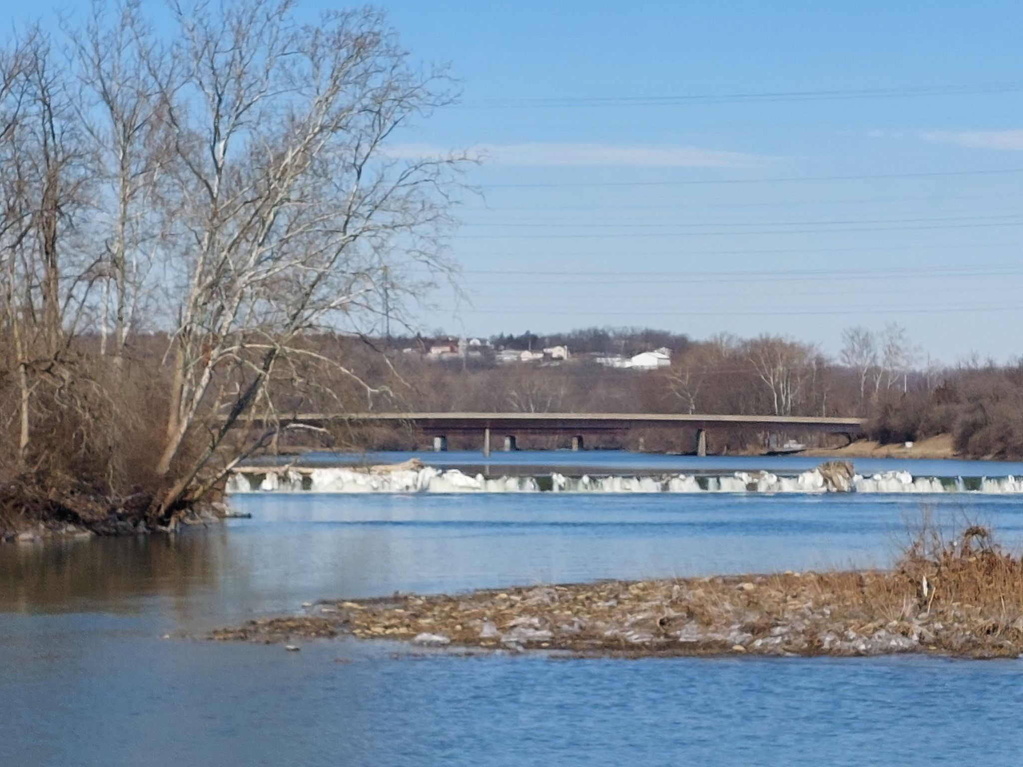 River with a low dam, distant bridge, and bare trees under a blue sky. - Hutchins State Reservoir loop from Chautauqua