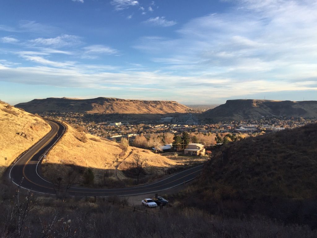 Winding road on golden hills overlooking a town and mountains under a blue sky. - Chimney Gulch Trail
