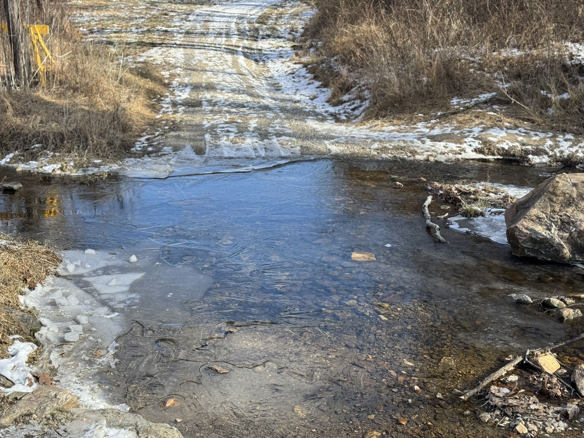 A shallow stream crossing a muddy track with patches of snow and dry grass. - Fair Hill Trailhead loop from Strickersville