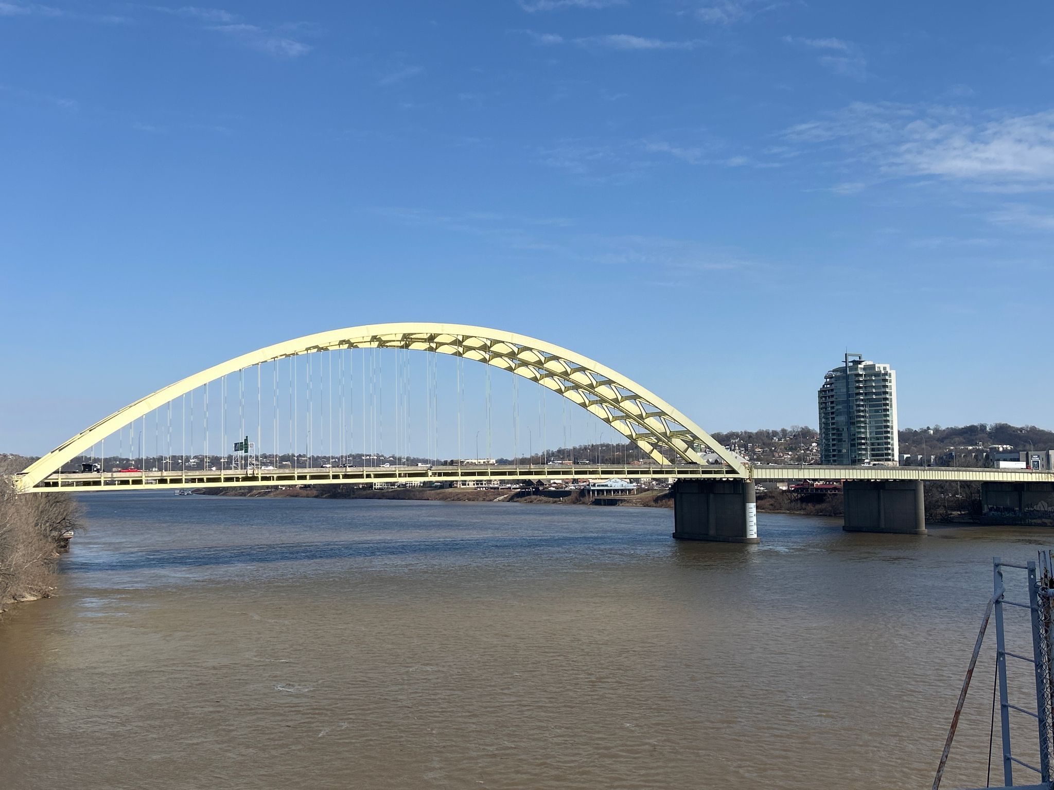 A large yellow arch bridge spans a wide river under a clear blue sky. - Cincinnati Flying Pig Statues – Ohio River Trail loop from Fort Thomas
