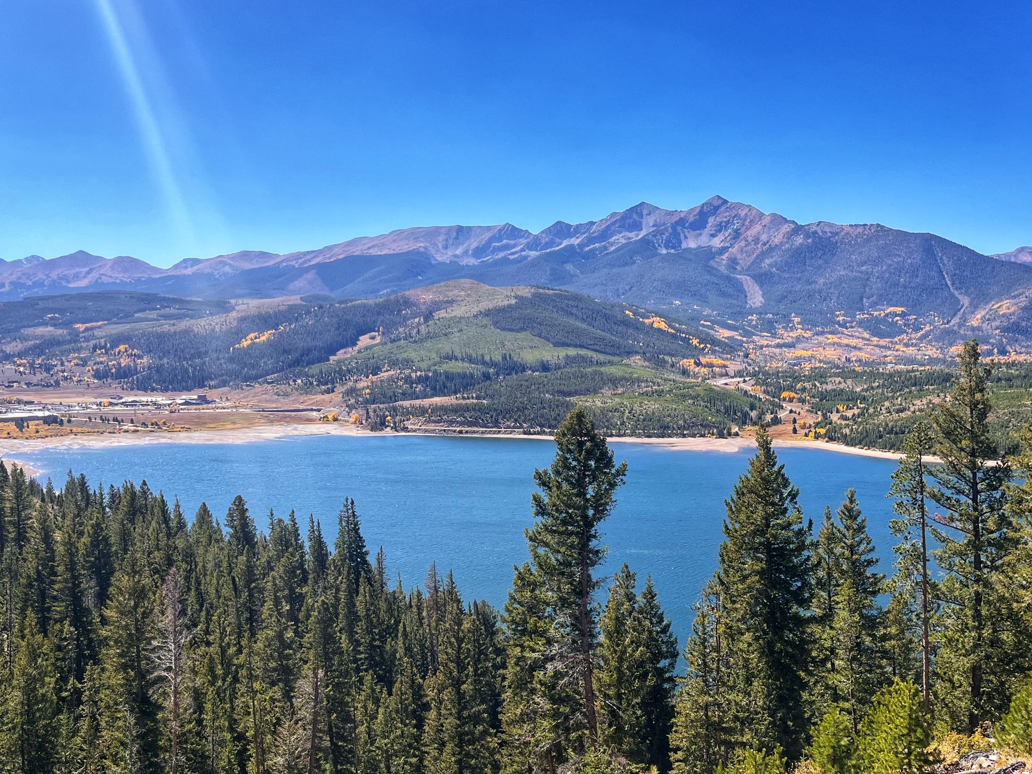 A bright sunny view of a large blue lake surrounded by forests and mountains. - Dillon Reservoir Recpath