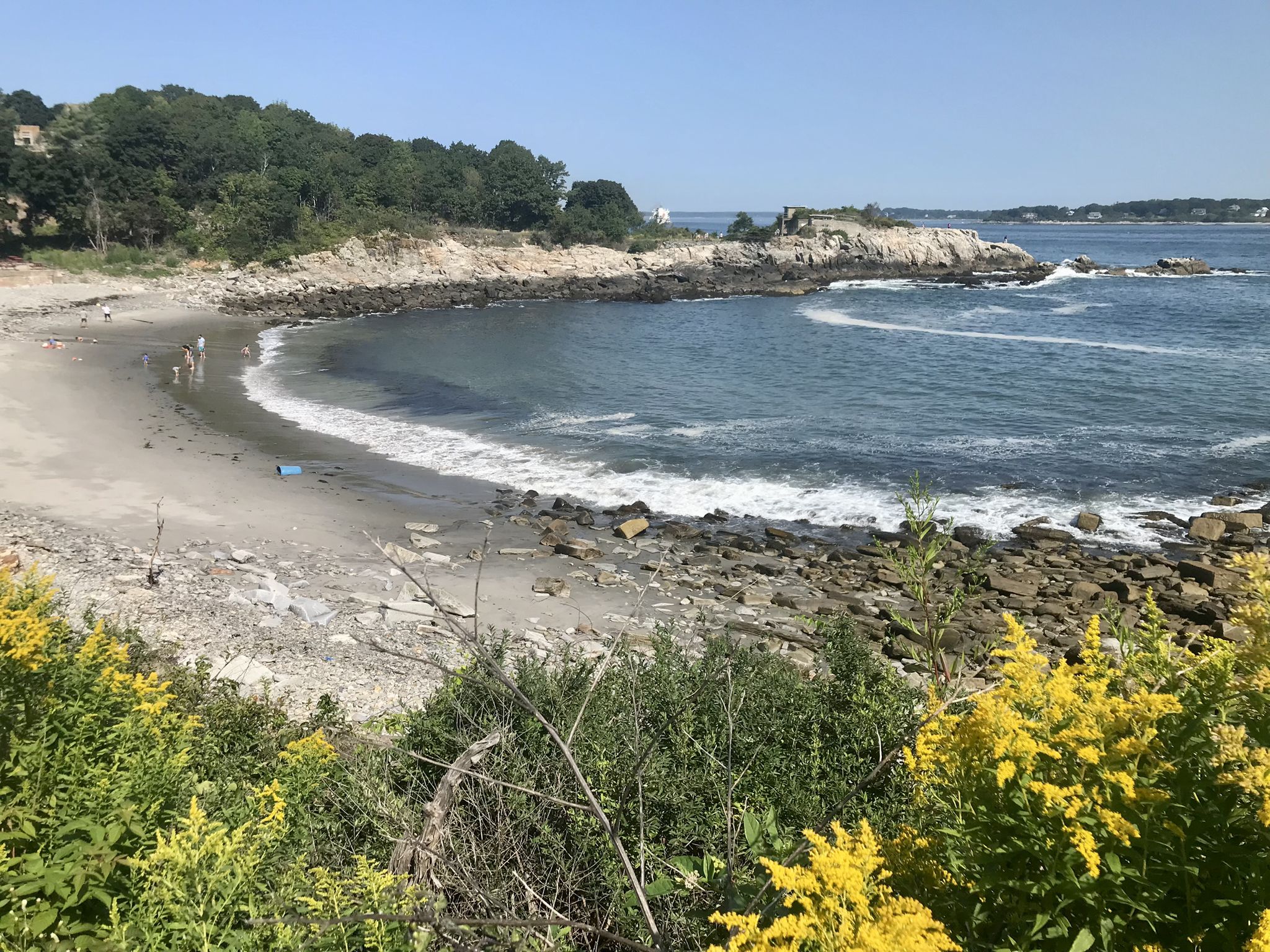 Sunny view of a curved sandy beach with waves, rocky coast, and people. - Fort Williams Bike Route