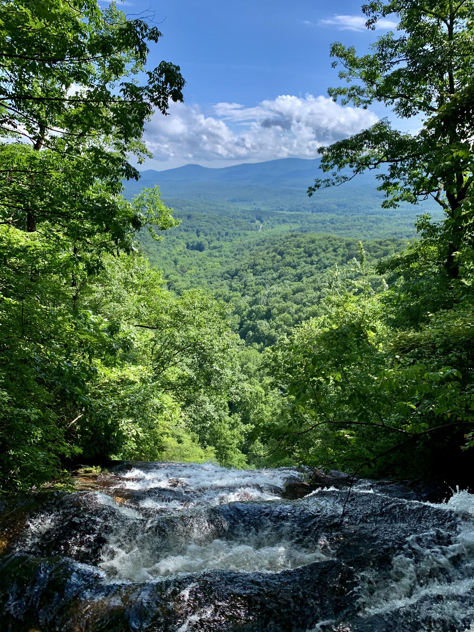 Waterfall cascading over rocks into a lush green forest valley under a blue sky. - A.T. Approach Trail to the Reflection Pool loop