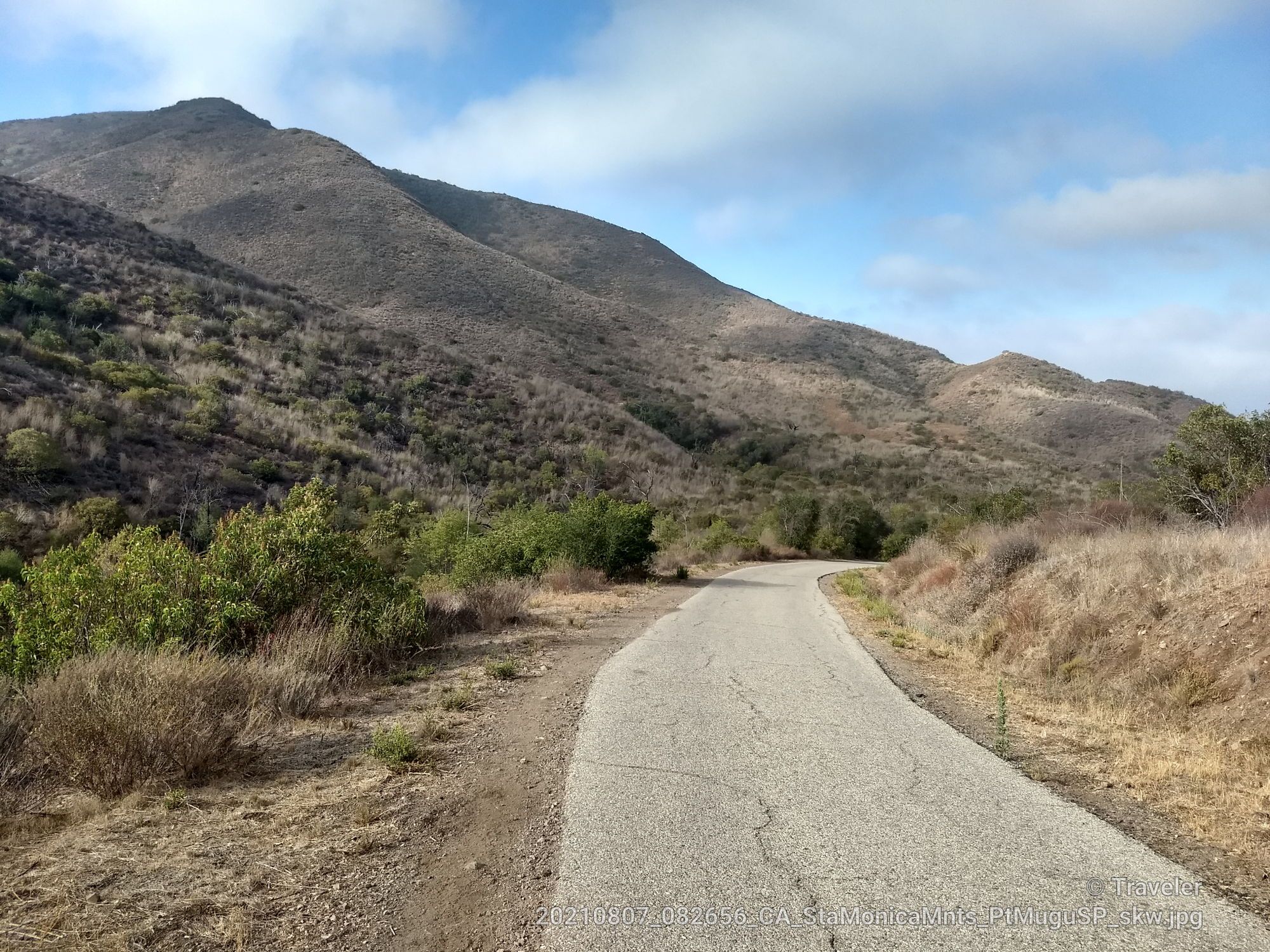 Paved road curves through dry, brush-covered hills under a partly cloudy sky. - Native Garden (trailheads) – Big Sycamore Canyon Summit loop from Rancho Sierra Vista/Satwiwa