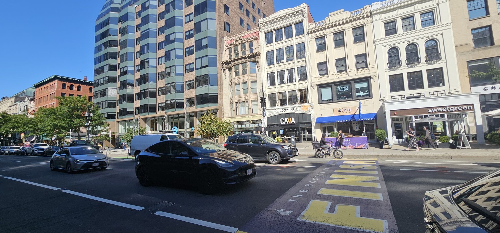 A sunny city street with cars, tall buildings, and pedestrians. - Boston Marathon Route