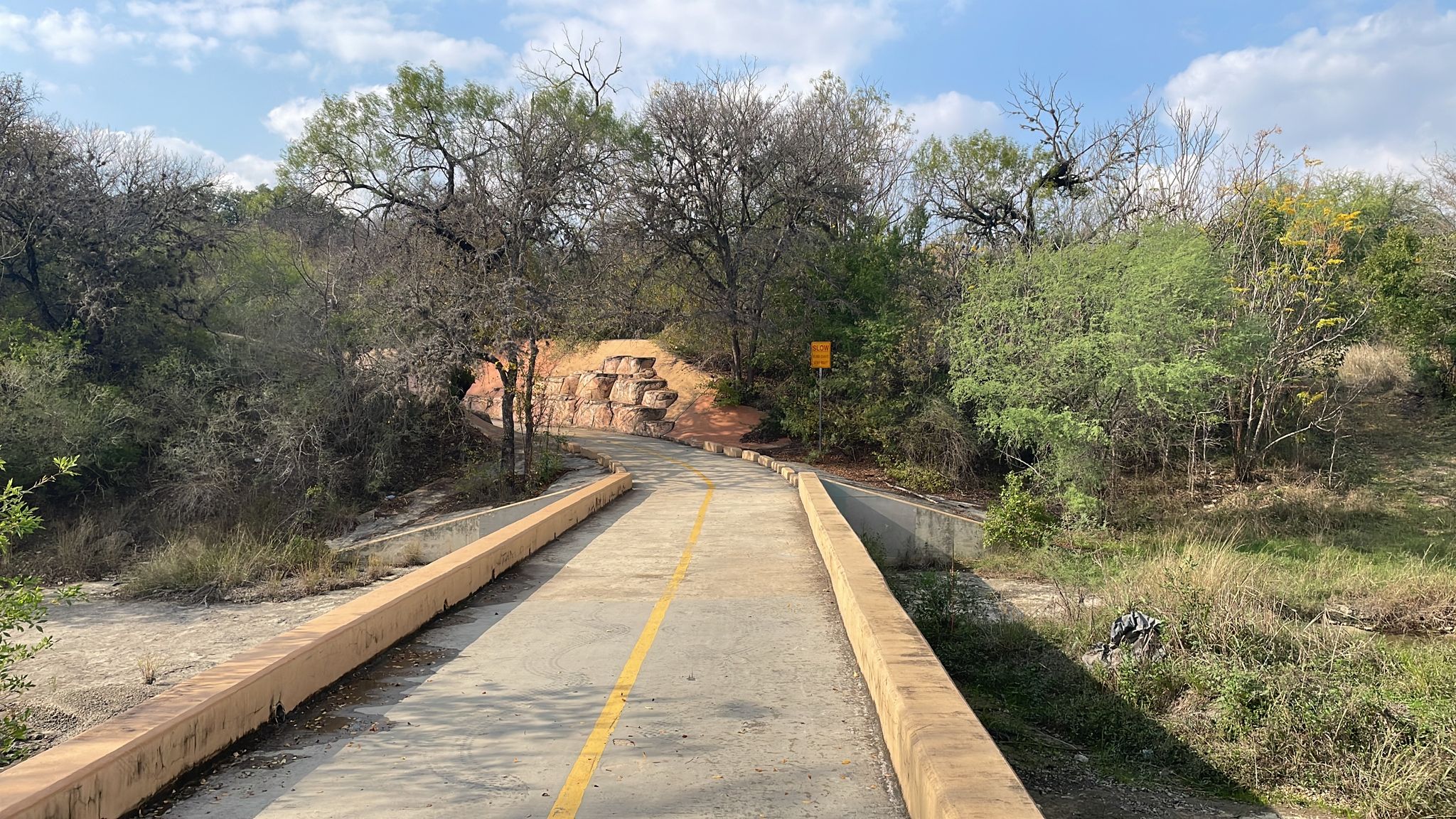A paved bridge with a yellow line leads into a wooded area under a cloudy sky. - Robert L.B. Tobin Park – Morningstar Boardwalk loop from McNay Art Museum