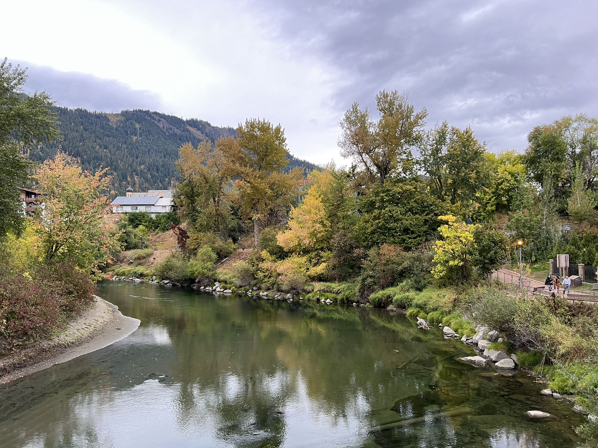 A river flows past autumn trees and distant mountains under a cloudy sky. - Barn Beach Pea Vine Trail