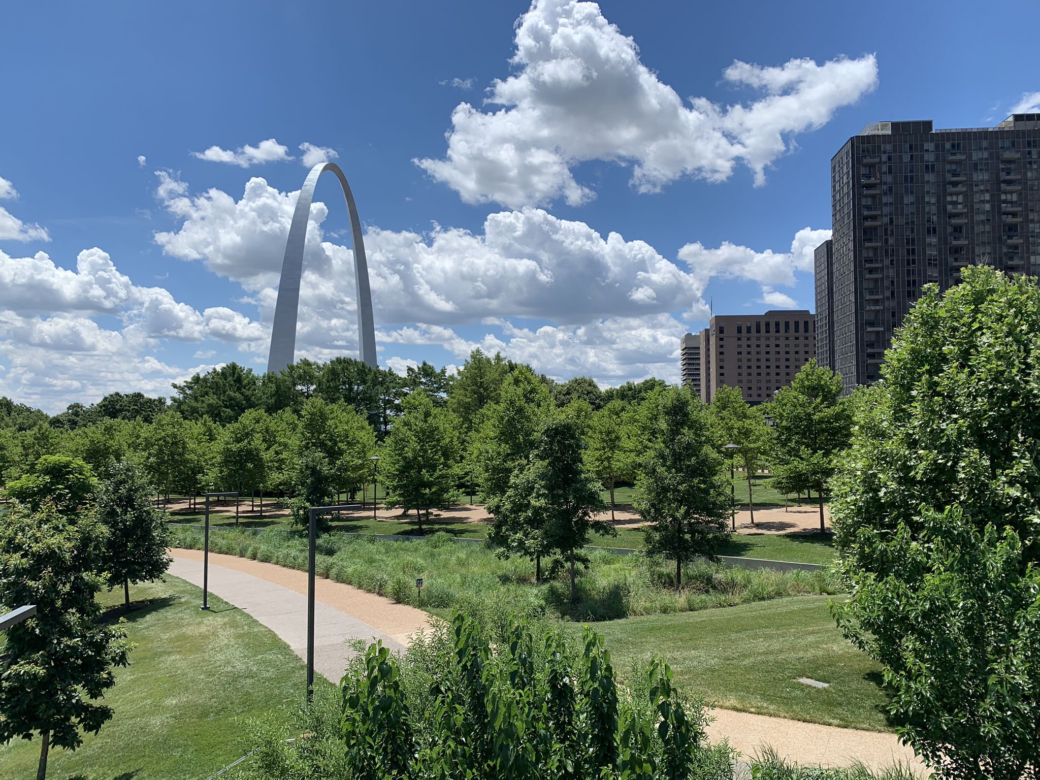 Large arch over a green park with trees and distant city buildings. - Gateway Arch and Riverfront Trail