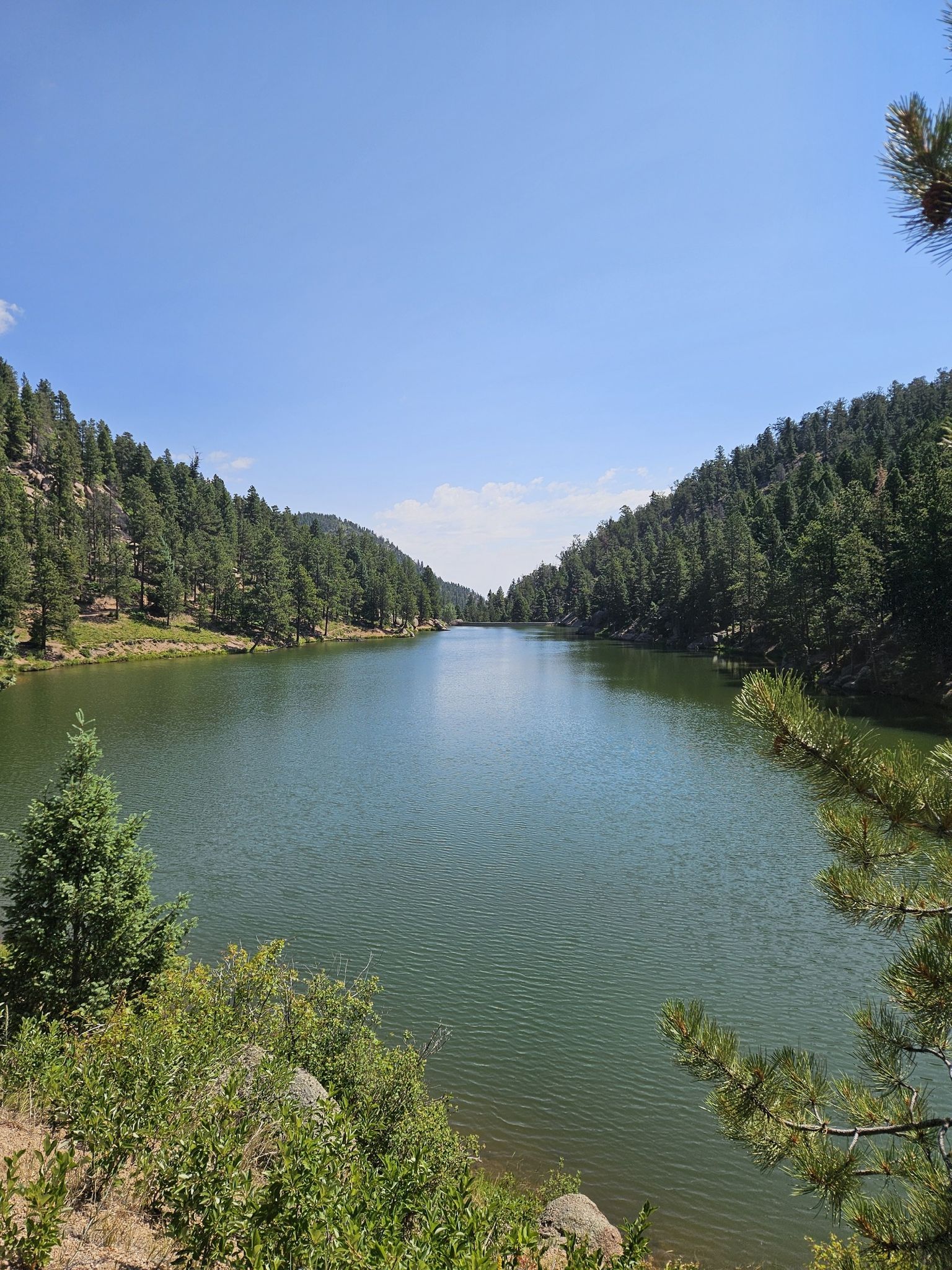 A serene lake surrounded by sunlit pine-covered hills under a clear blue sky. - La Deux Réservoirs Trail