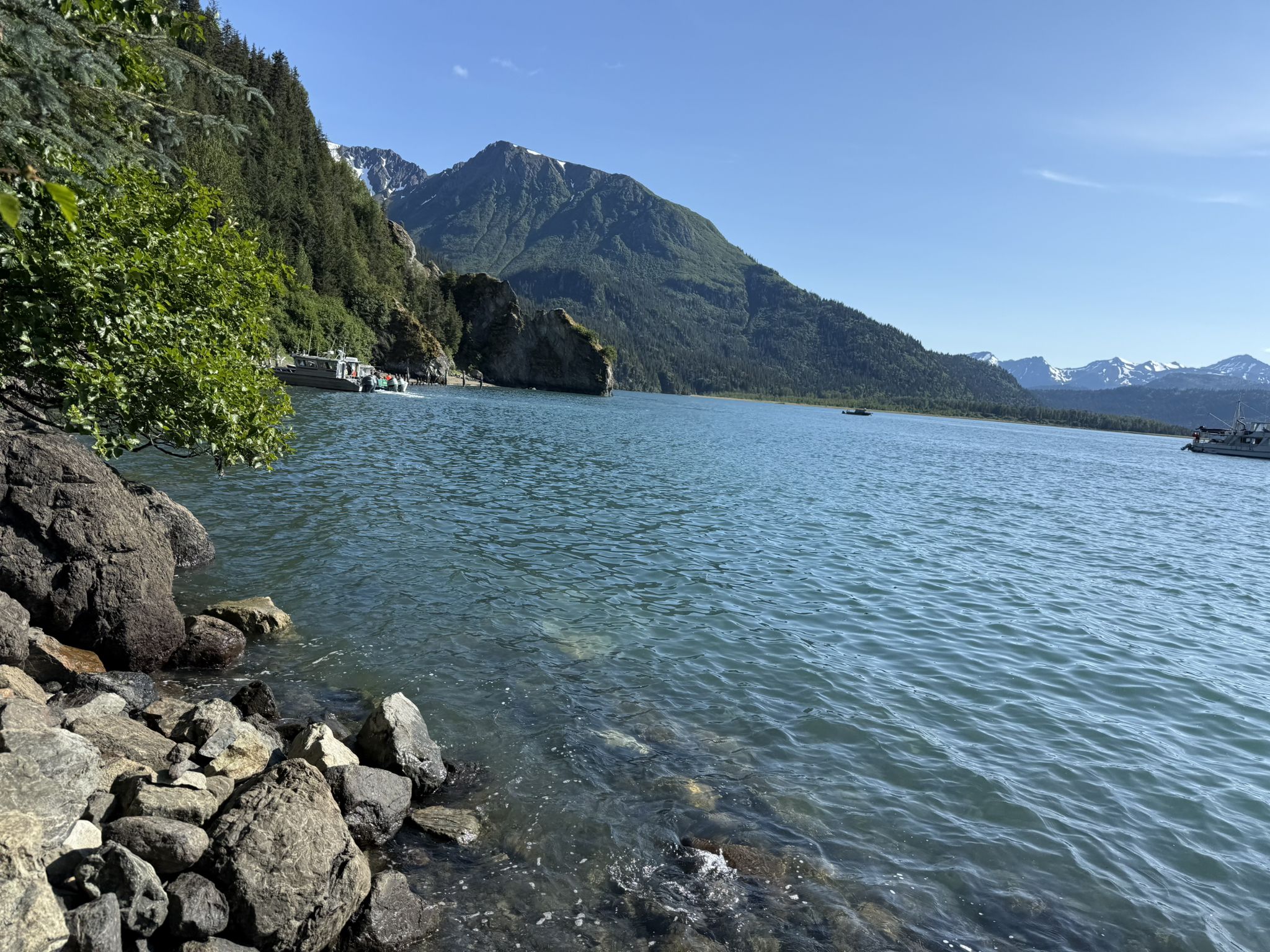 Clear blue water with rocky shore, forested mountains, and distant boats. - Saddle Trail to Grewingk Lake
