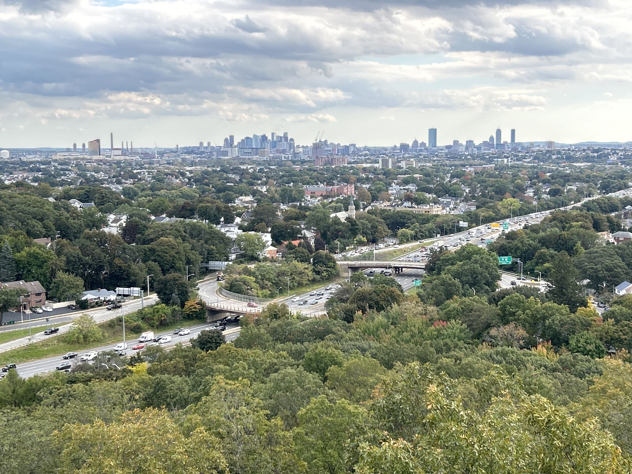 Distant city skyline over green trees and a busy highway under cloudy skies. - Southern Middlesex Fells Loop