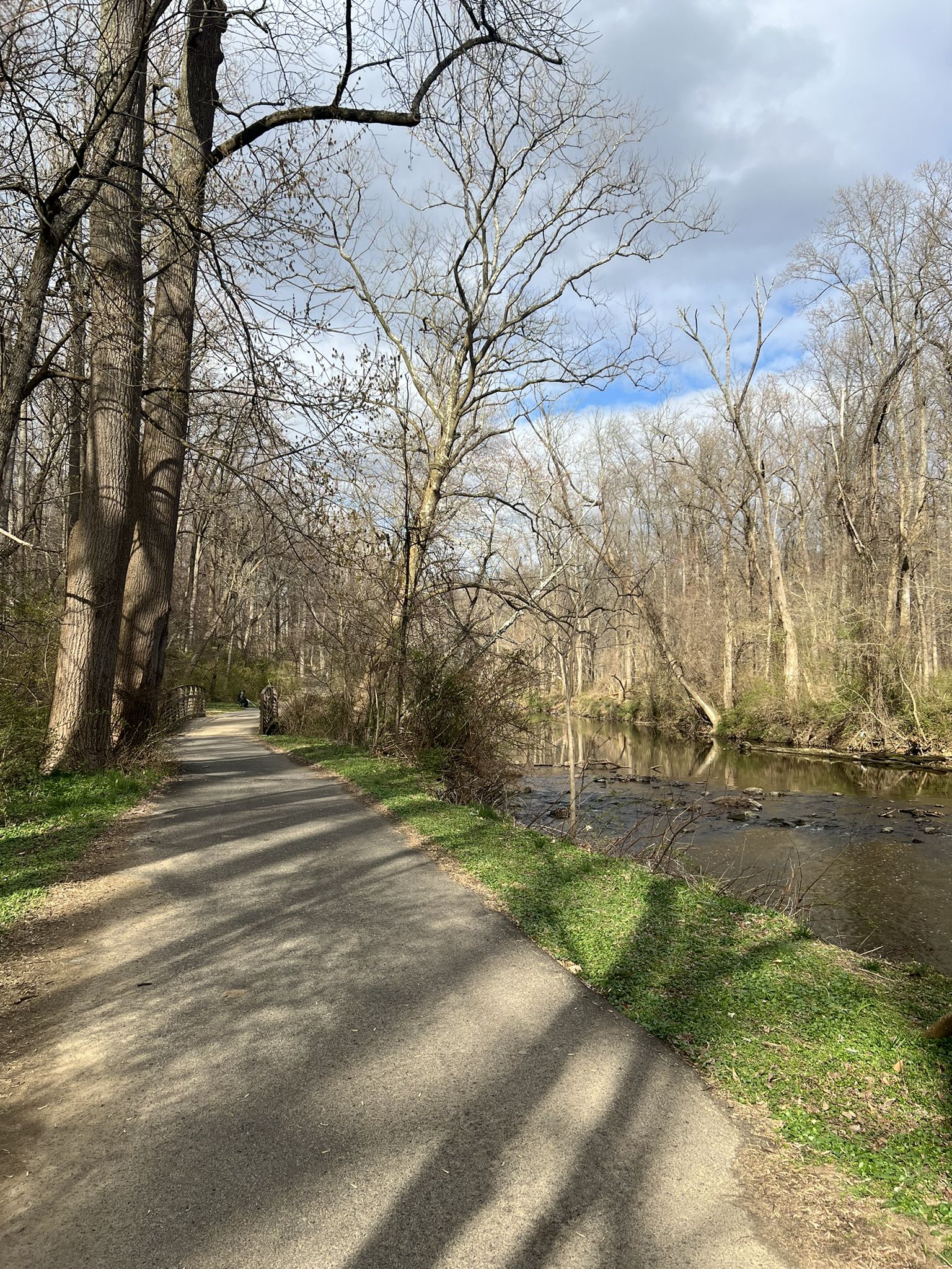 A paved path runs alongside a river in a wooded area under a cloudy sky. - Pennypack Trail Loop