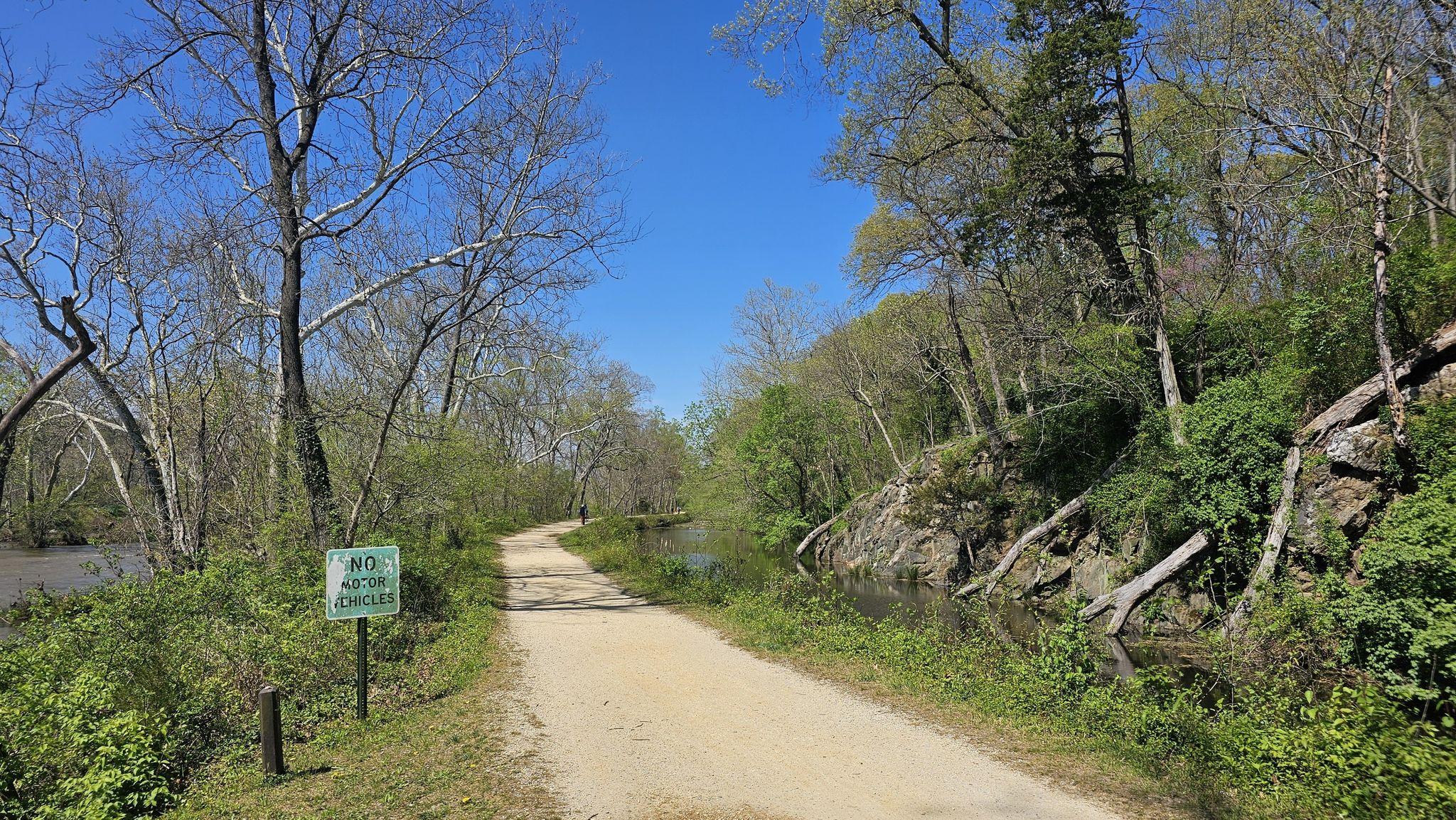 A sunny dirt path runs beside a canal and wooded hill. - Francis Scott Key Bridge – Crossing Over the C&O Canal loop from Potomac