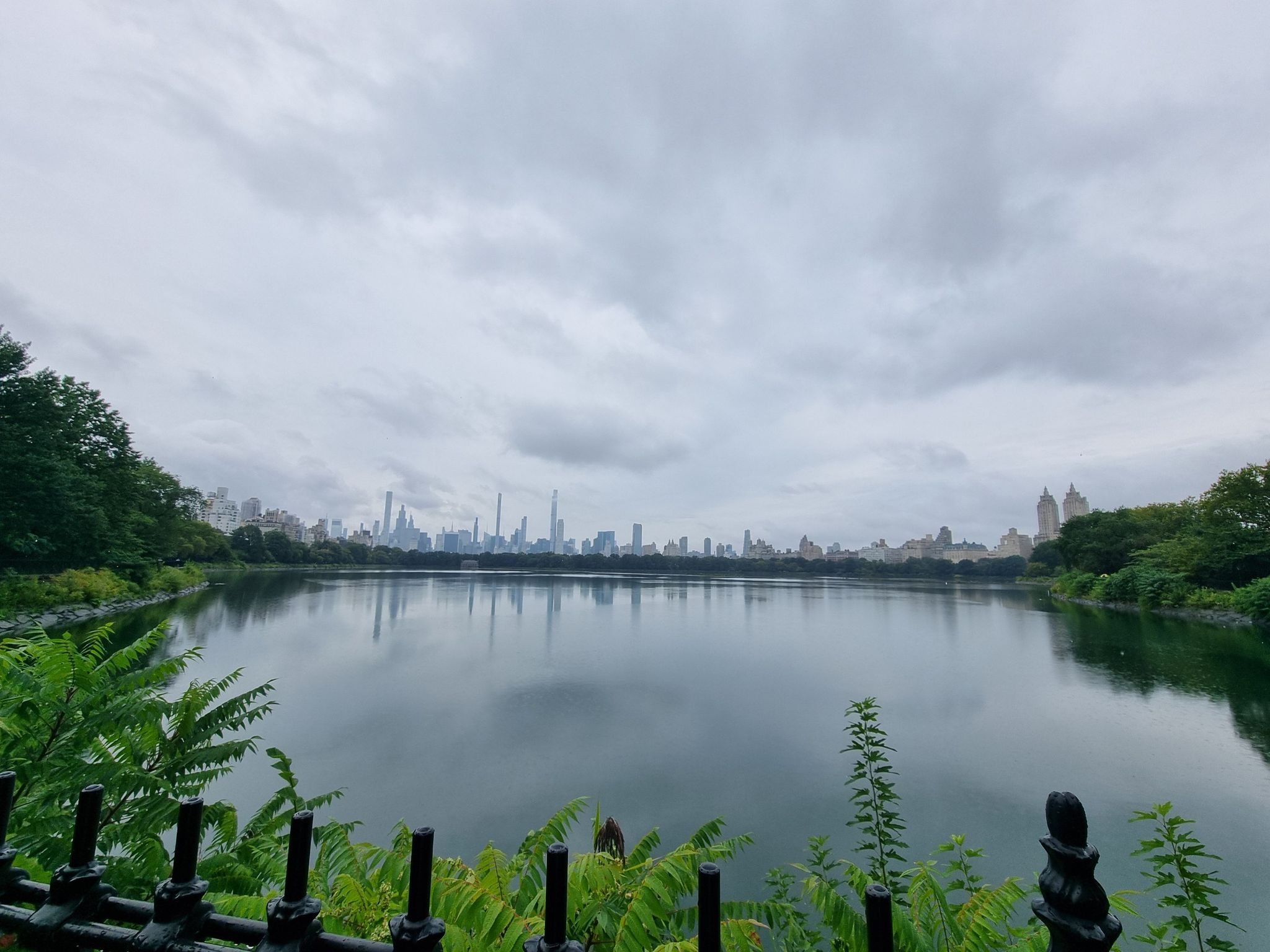 Lake reflecting a distant city skyline under a cloudy sky. - View of the San Remo Towers – Bethesda Fountain and Terrace loop from 66th Street–Lincoln Center