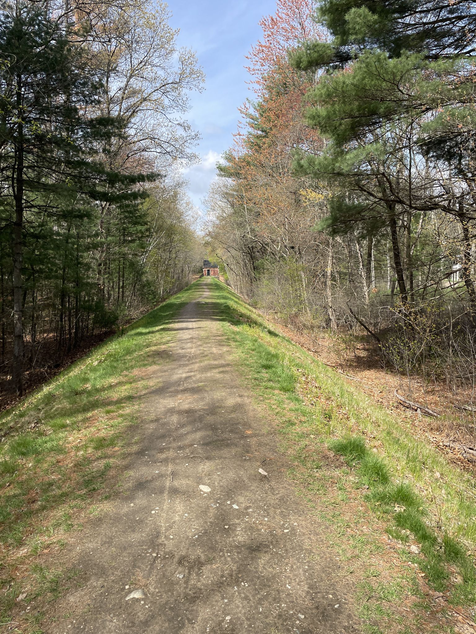 Dirt path through a forest, leading to a distant building under bright sky. - Echo Bridge – Sudbury Aqueduct Path loop from Boulder Brook Reservation