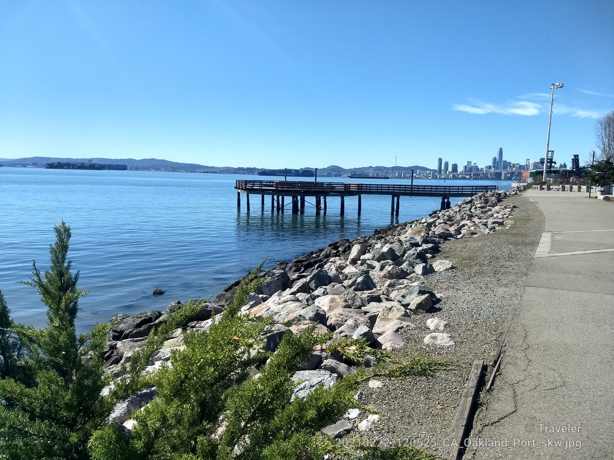 Calm bay with a long pier, rocky shore, and distant city skyline under blue sky. - San Francisco Bay Trail: Oakland and Treasure Island Loop