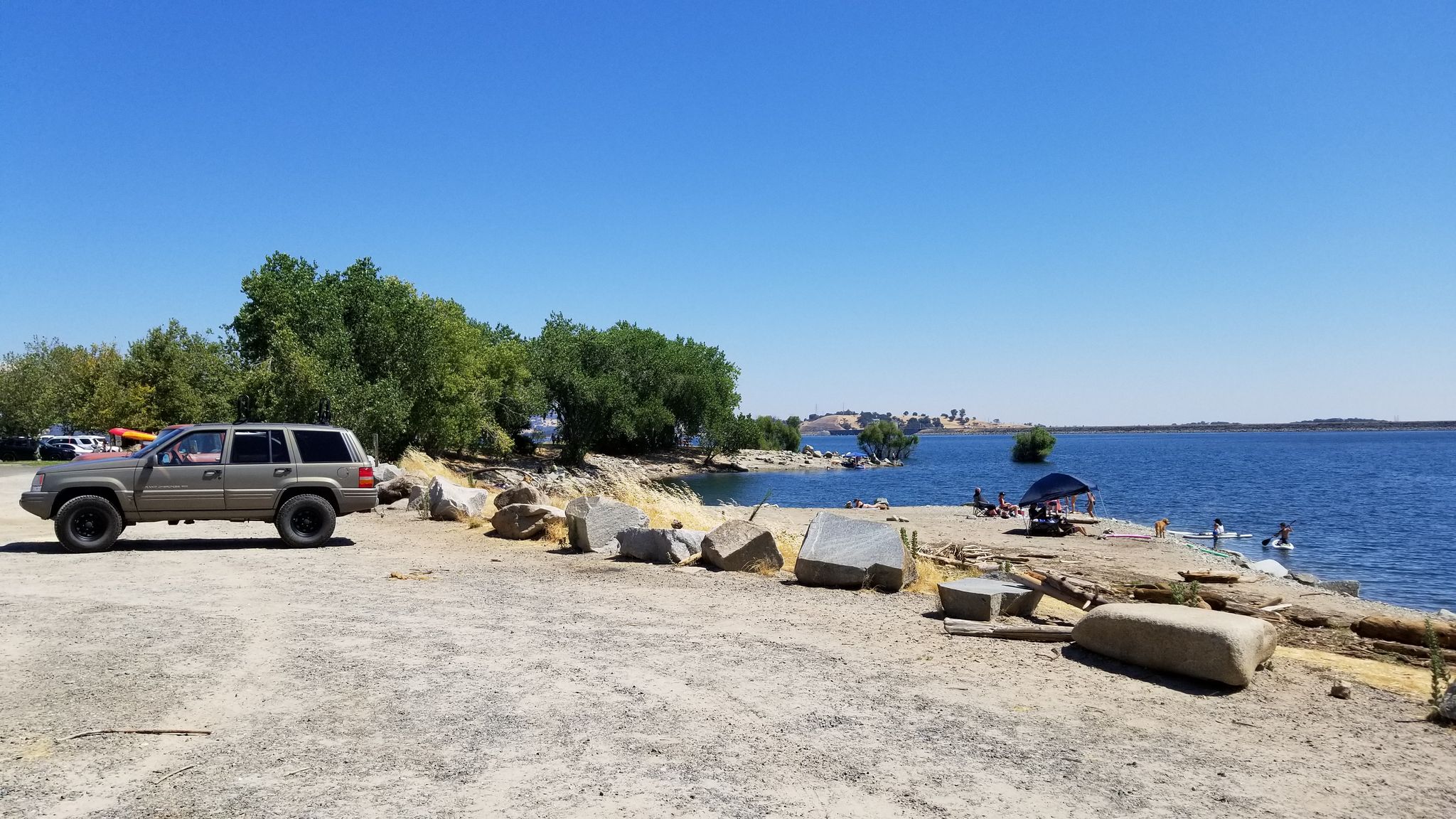 An SUV parked by a lake with people swimming on a sunny day. - American River Bike Trail