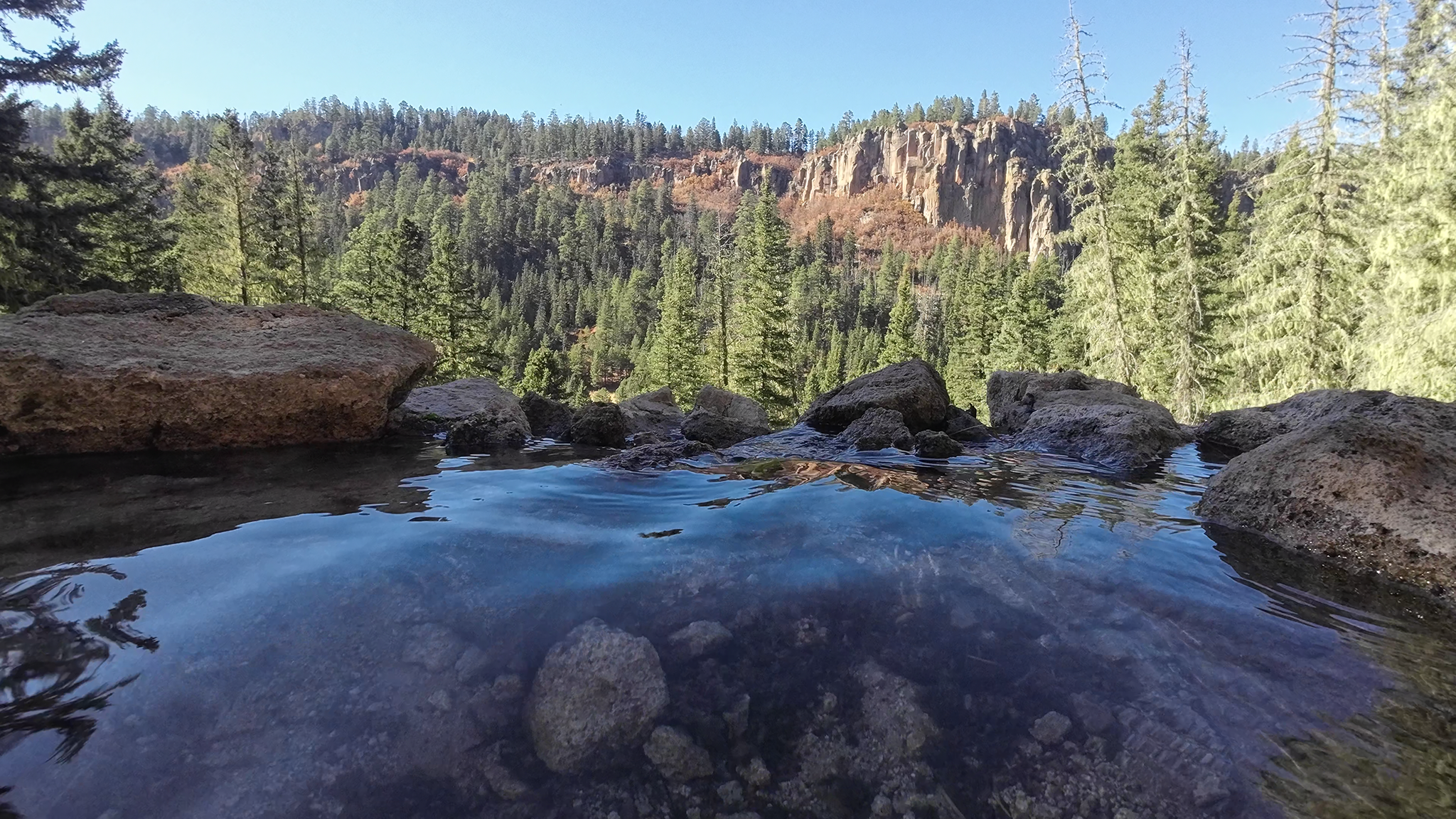 Clear water pool with rocks, pine forest, and sunny cliff face. - San Antonio Hot Springs Trail
