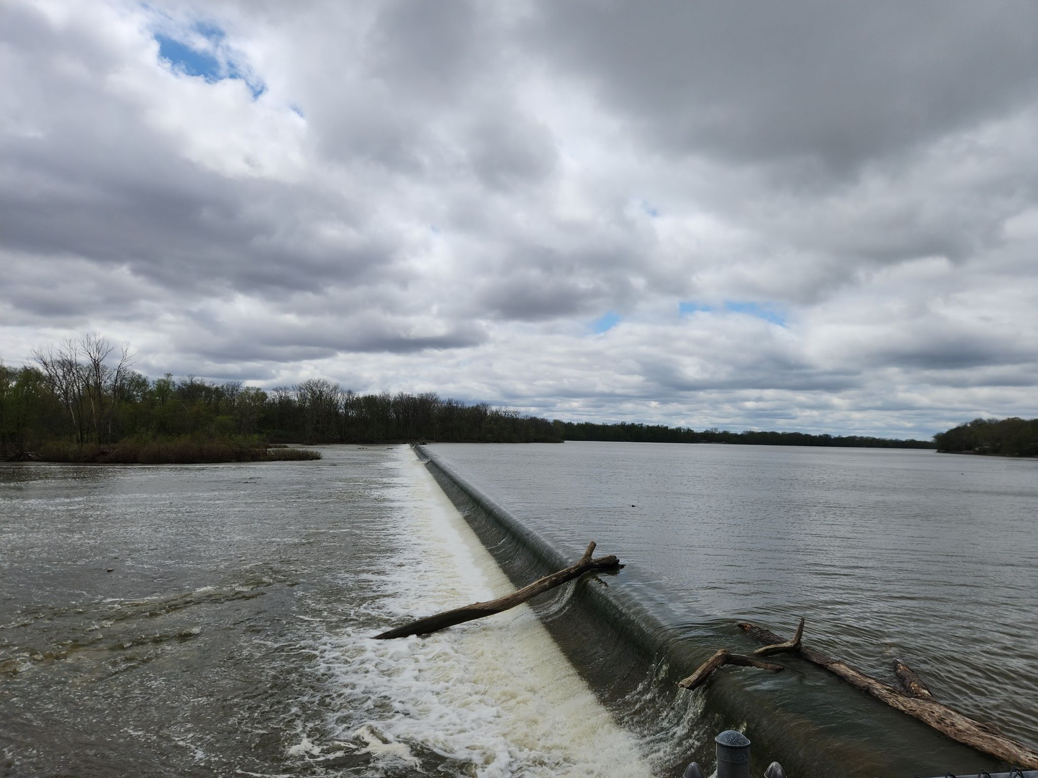 A wide river flows over a low dam under a cloudy sky. - Towpath Trail