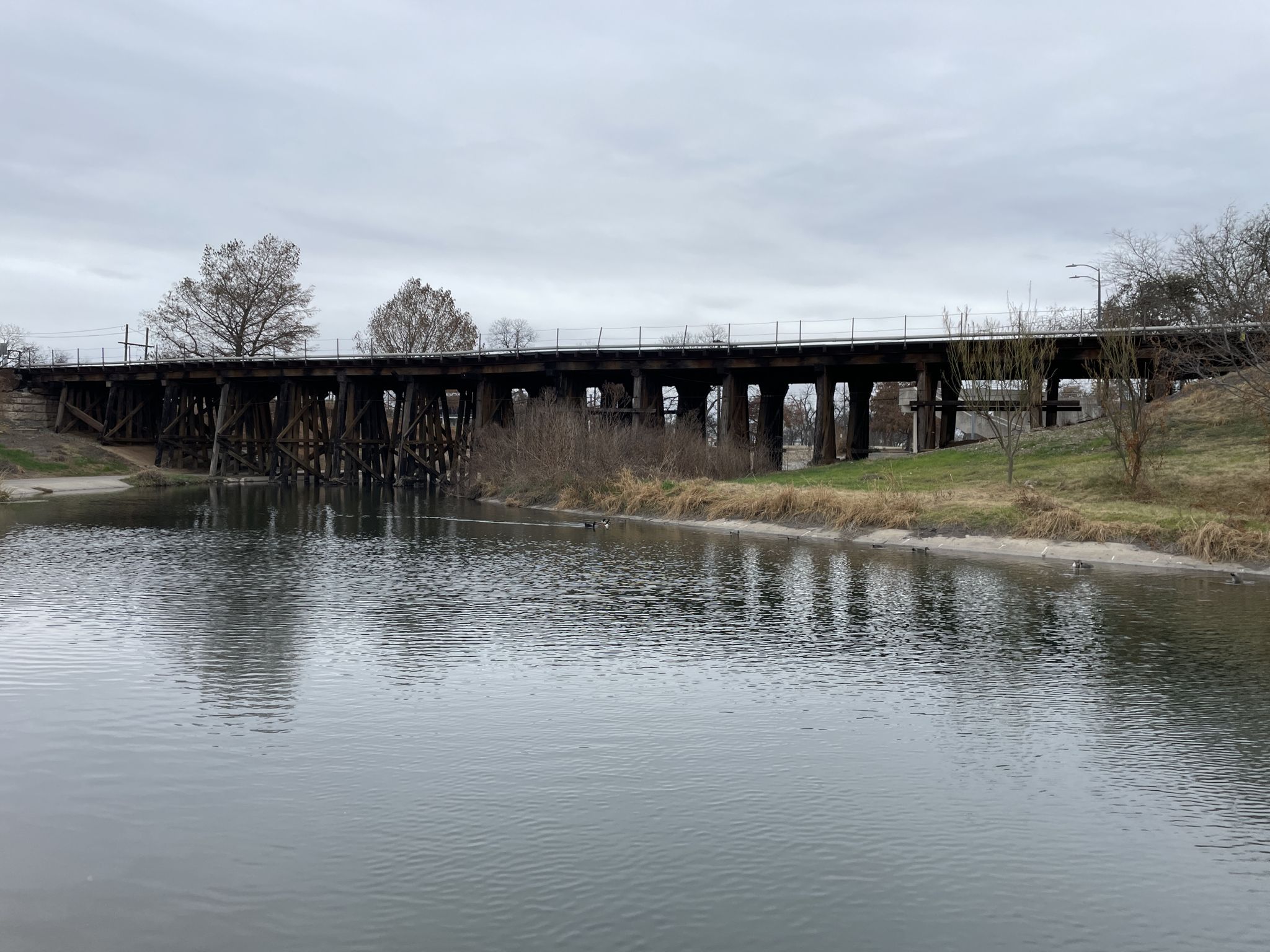 Long wooden train bridge over a calm river under an overcast sky. - Alamo Mills Dam – Hays Street Bridge loop from Olmos Park