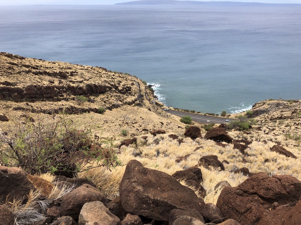 Rocky, dry hillside overlooking a coastal road and ocean under an overcast sky. - Lahaina Pali Trail