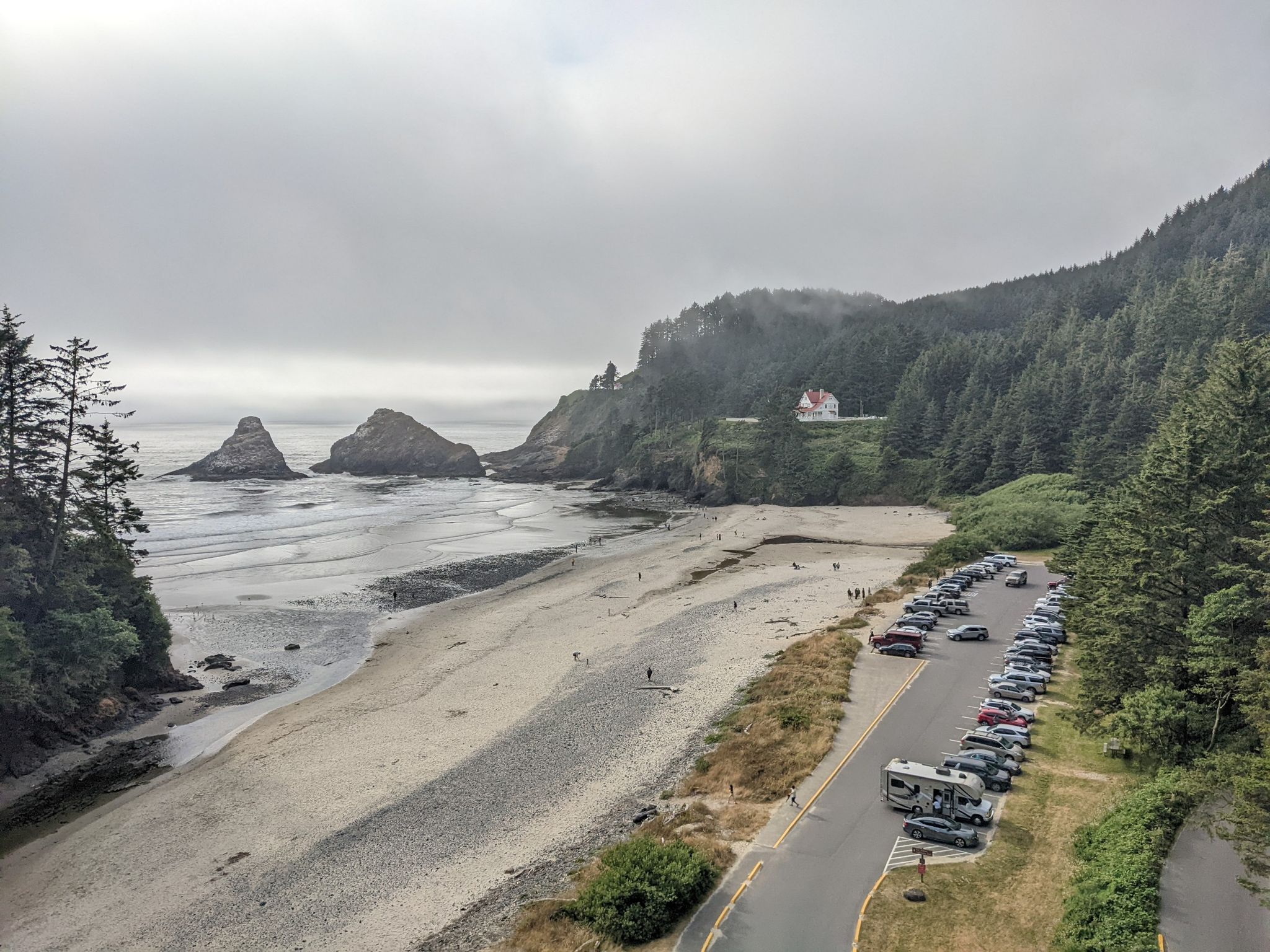 Overcast view of a sandy beach with sea stacks, coastal forest, and parked cars. - Oregon Coast Scenic Bikeway