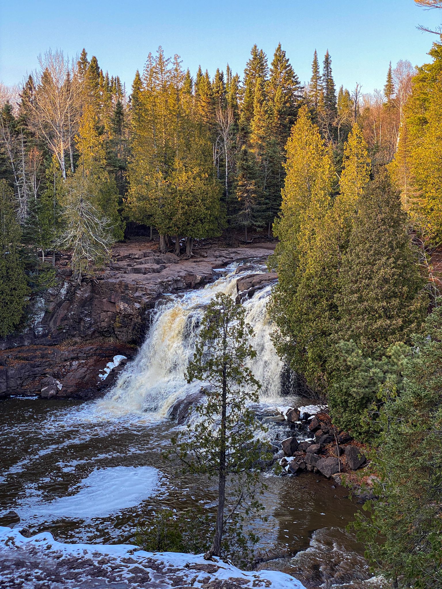 Waterfall cascades into a river, surrounded by sunlit evergreen and bare trees. - Fifth Falls – Upper Falls loop from Gooseberry Falls State Park