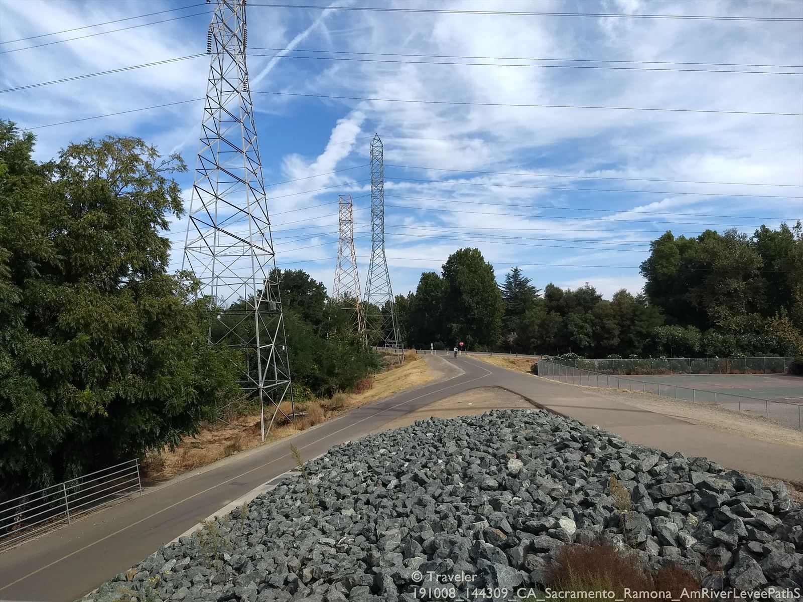 Paved path next to rocky embankment with power lines overhead on a bright day. - American River Levee Path