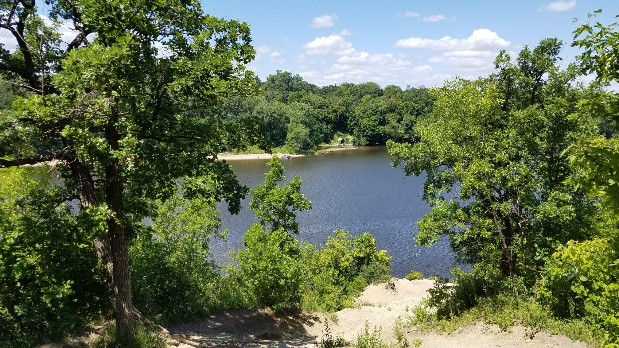 Sunny view of a calm lake surrounded by dense green trees. - Summit Ave Bikeway