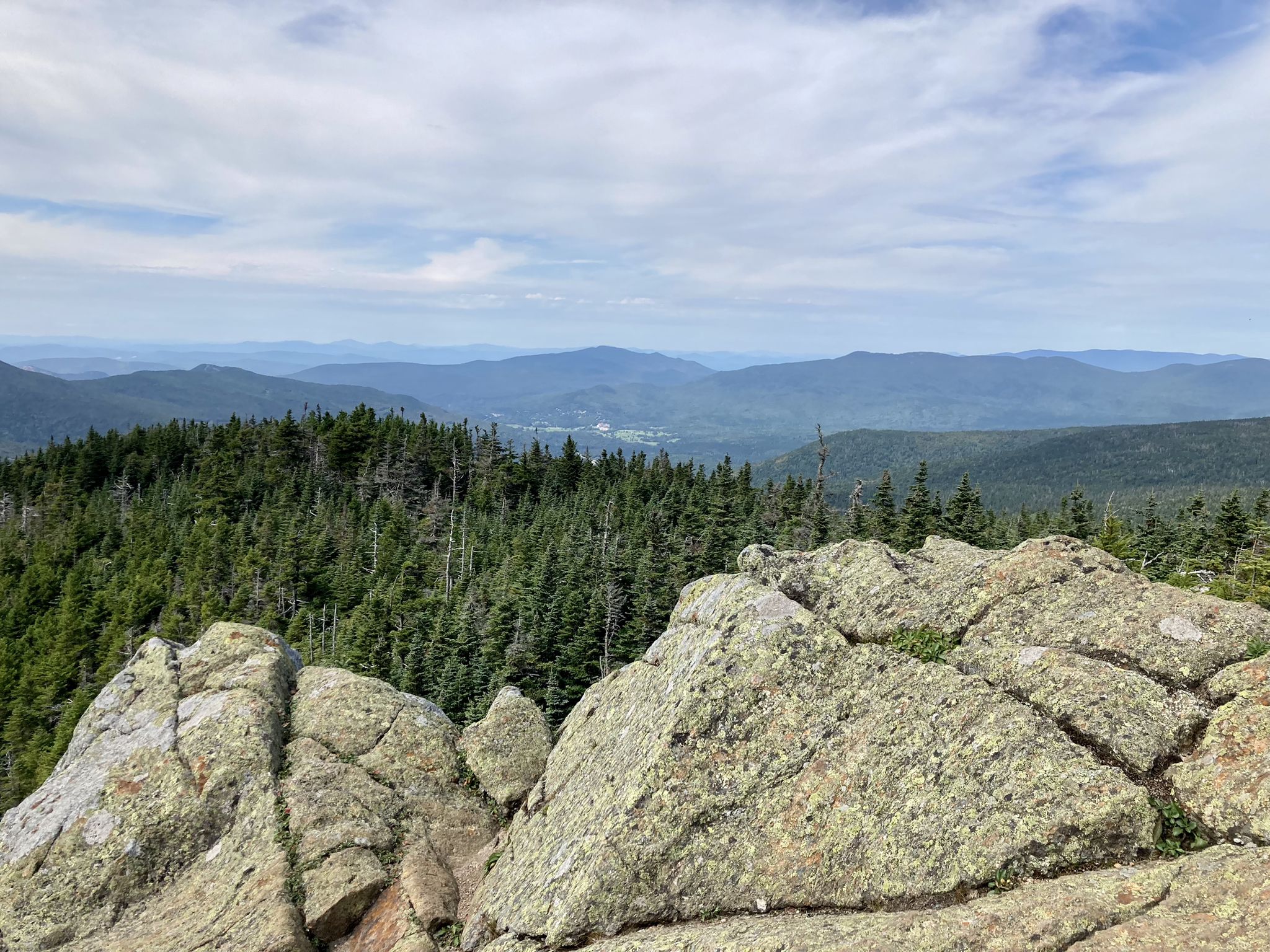 Rocky foreground overlooks dense forest, distant mountains, and a valley under a cloudy sky. - Presidential Traverse trail run