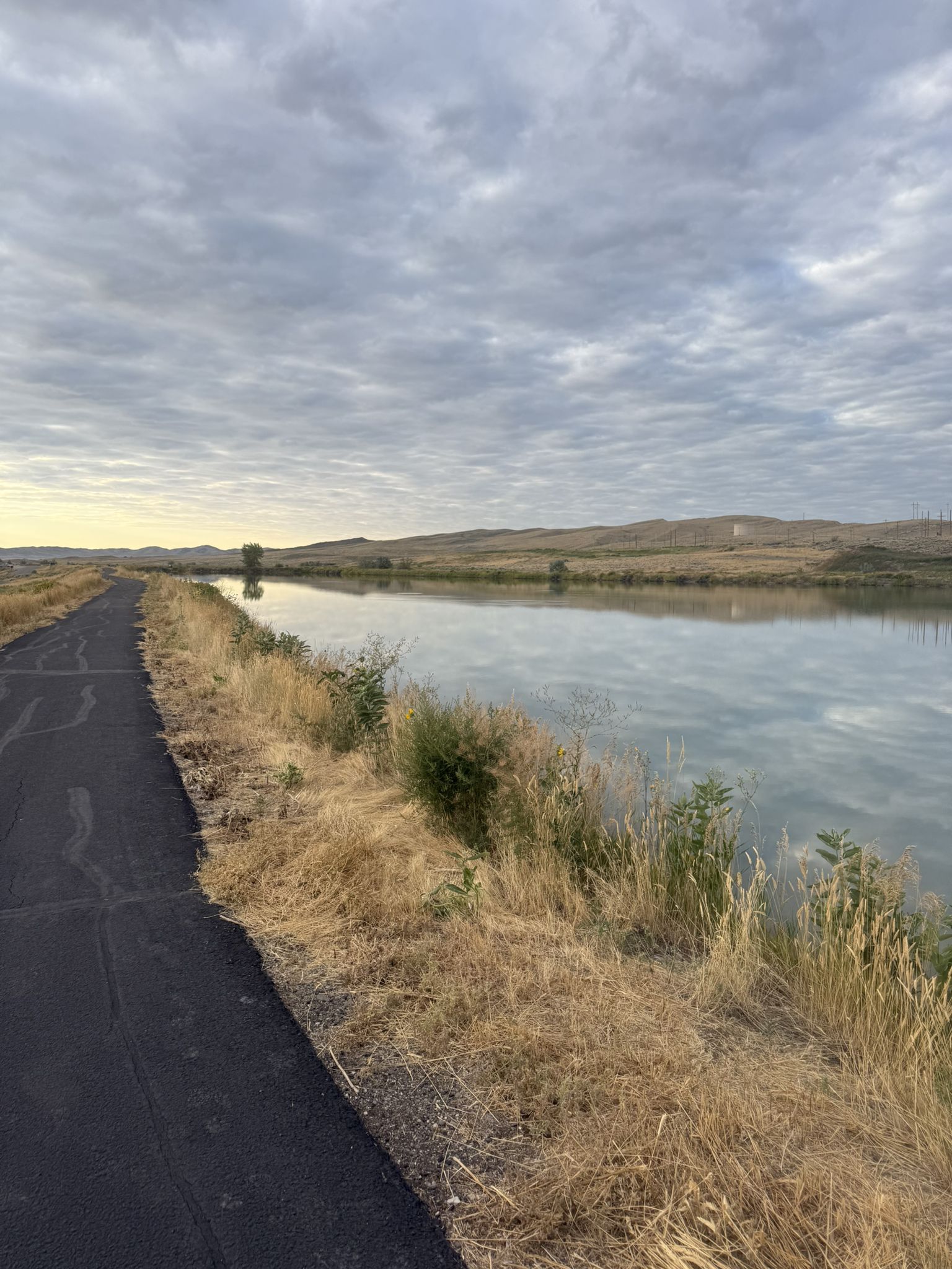 Paved path next to a calm canal with dry grass and distant hills. - New Cody Reservoir loop from Cody