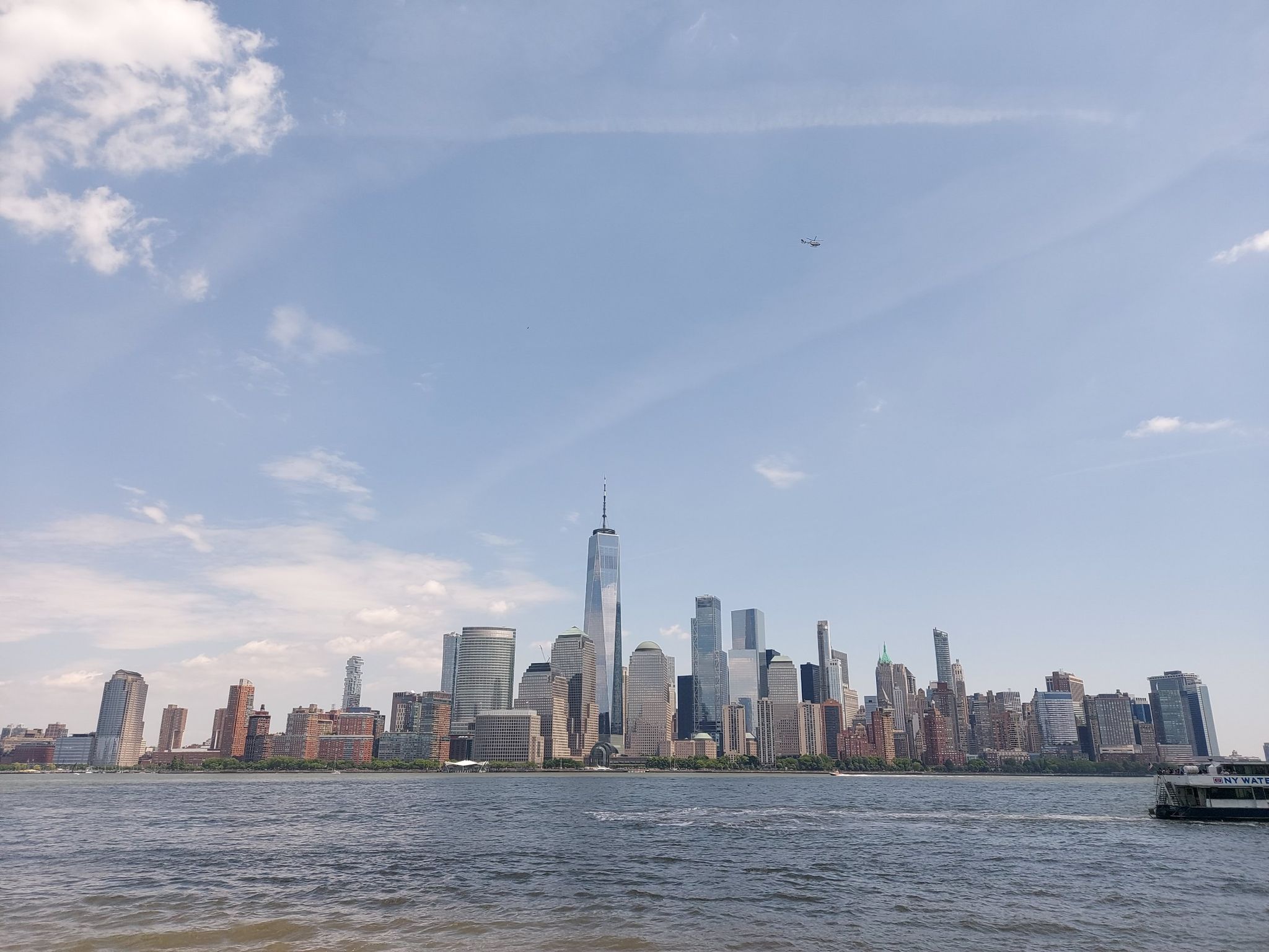 City skyline across water with a boat, under a bright, partly cloudy sky. - Jersey City 9/11 Memorial – View of the Manhattan Skyline loop from Harsimus Cove