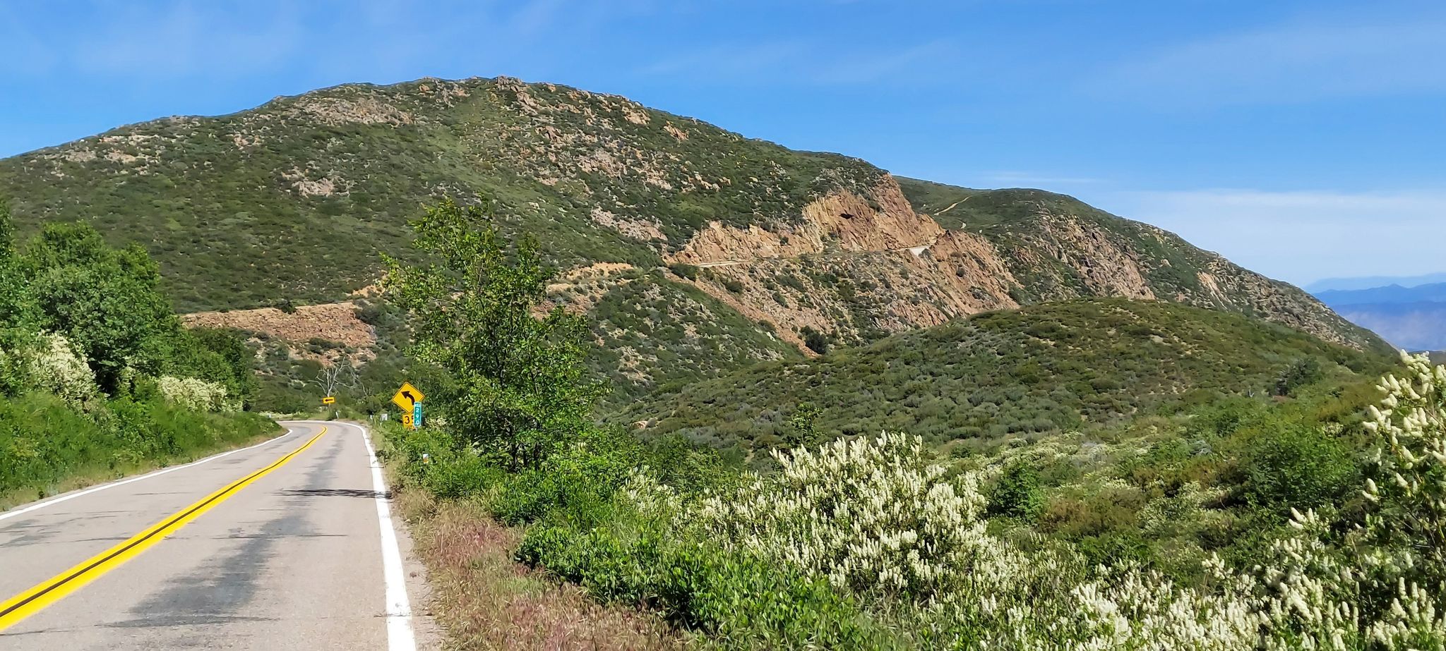 A paved road winds past a green, rocky mountain under a clear, sunny sky. - Garnet Peak Trail