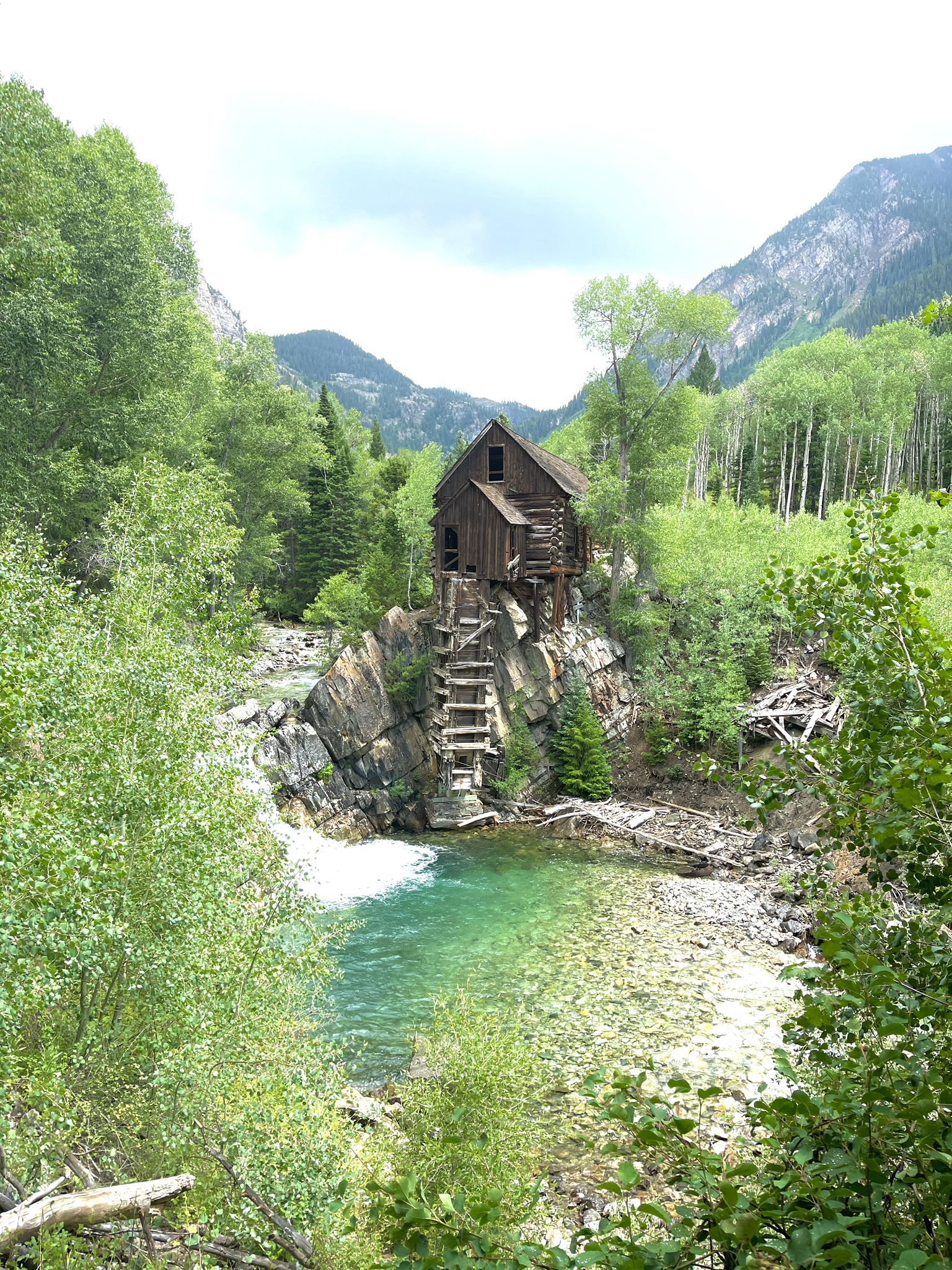 Rustic wooden building on a rocky outcrop overlooking a clear river, surrounded by green trees. - Crystal City Road #314