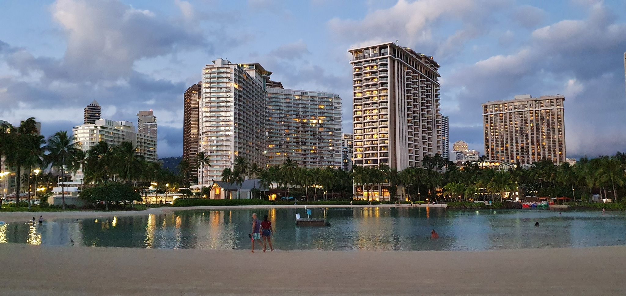 Illuminated city buildings reflecting on a calm lagoon at dusk with people. - Duke Kahanamoku Lagoon – Waikiki Beach loop from Diamond Head