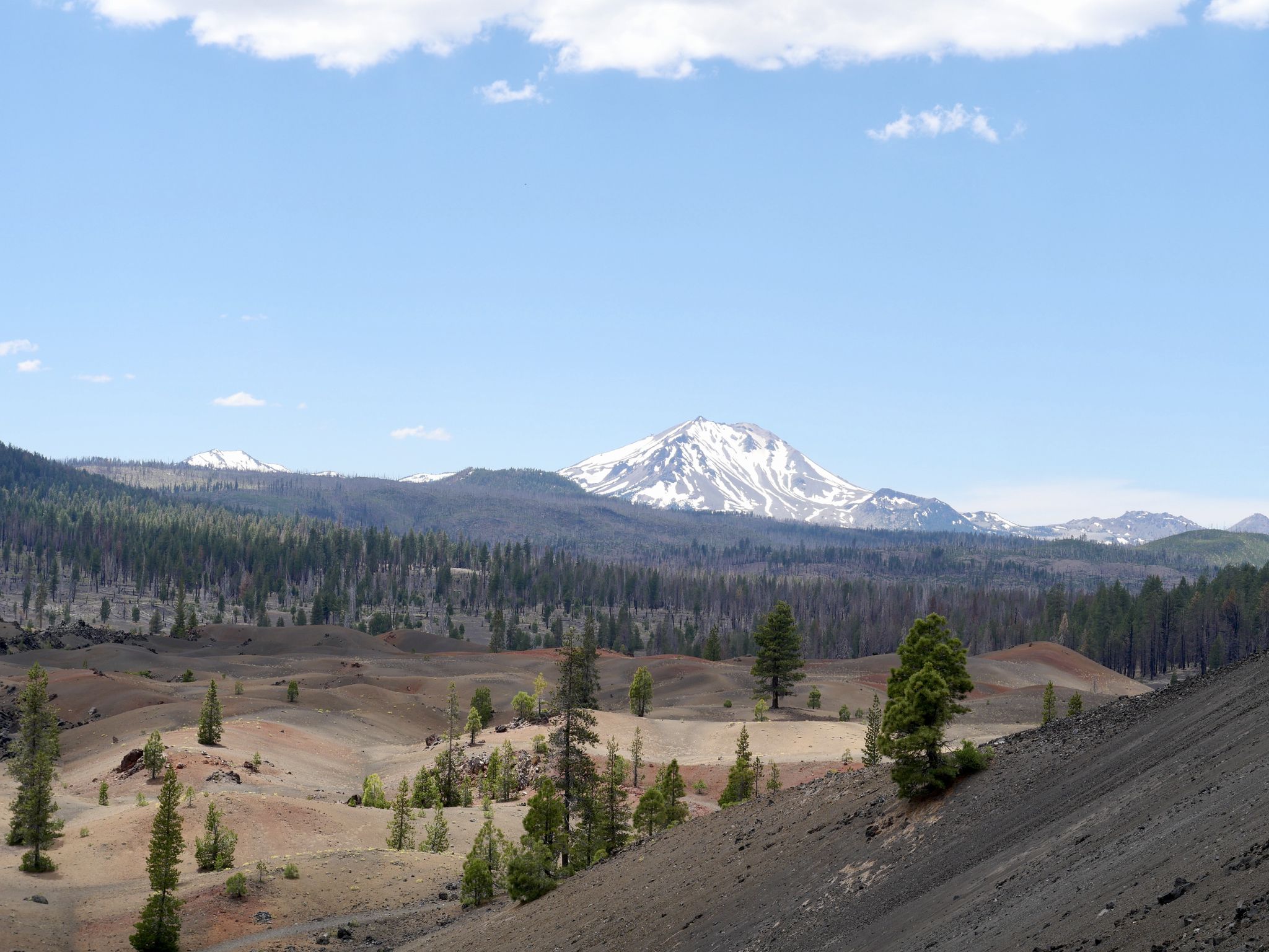 Distant snow-capped mountain overlooks a forested valley with sandy volcanic terrain. - Cinder Cone Trail