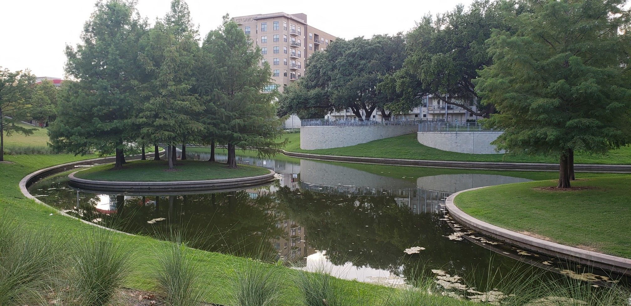Serene pond with a tree-covered island, green banks, and a distant building. - Running loop from Savoye