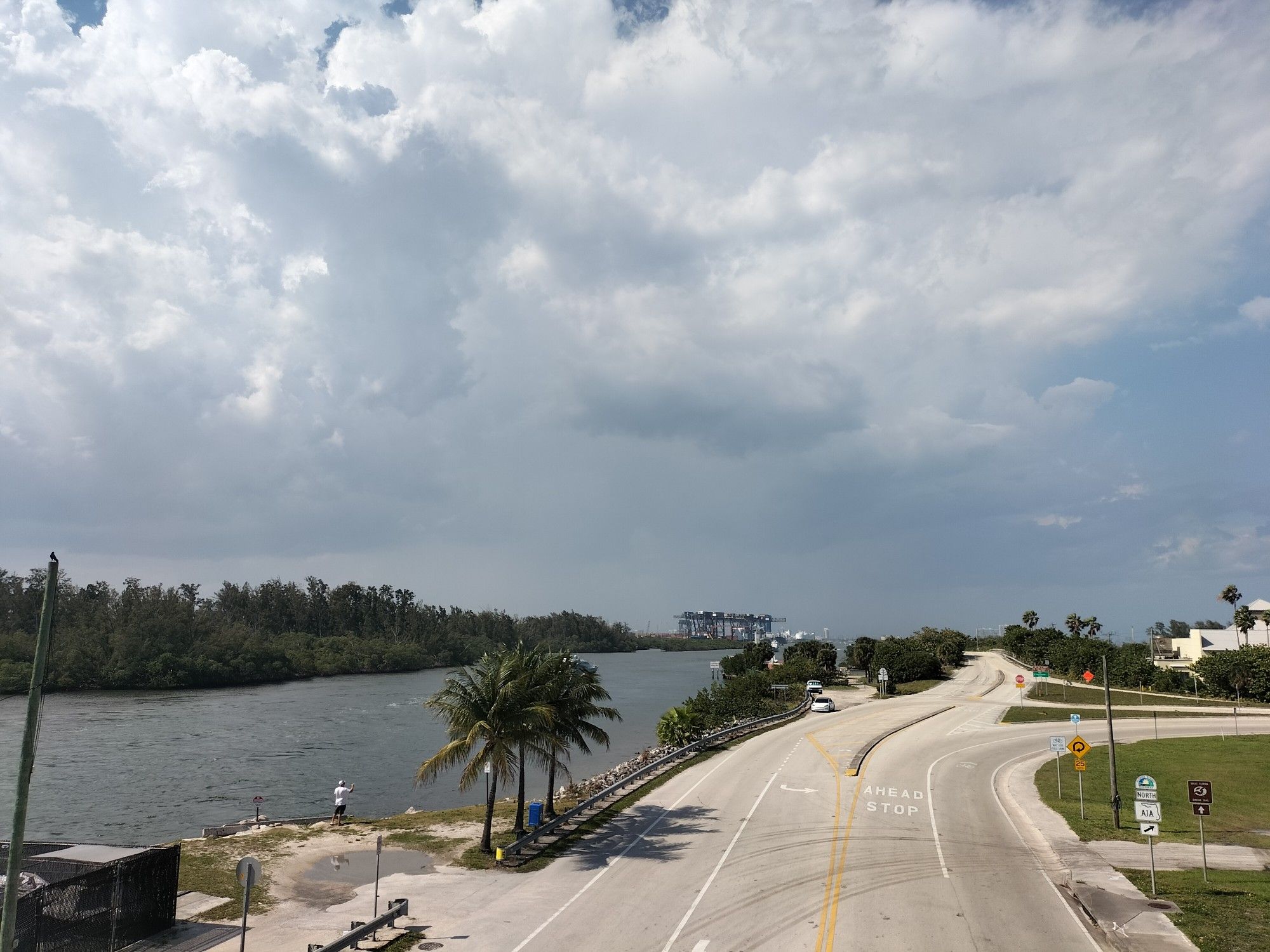 Overcast sky above a curving road, river, and distant industrial cranes. - Dania Beach Fishing Pier loop from Dania Beach