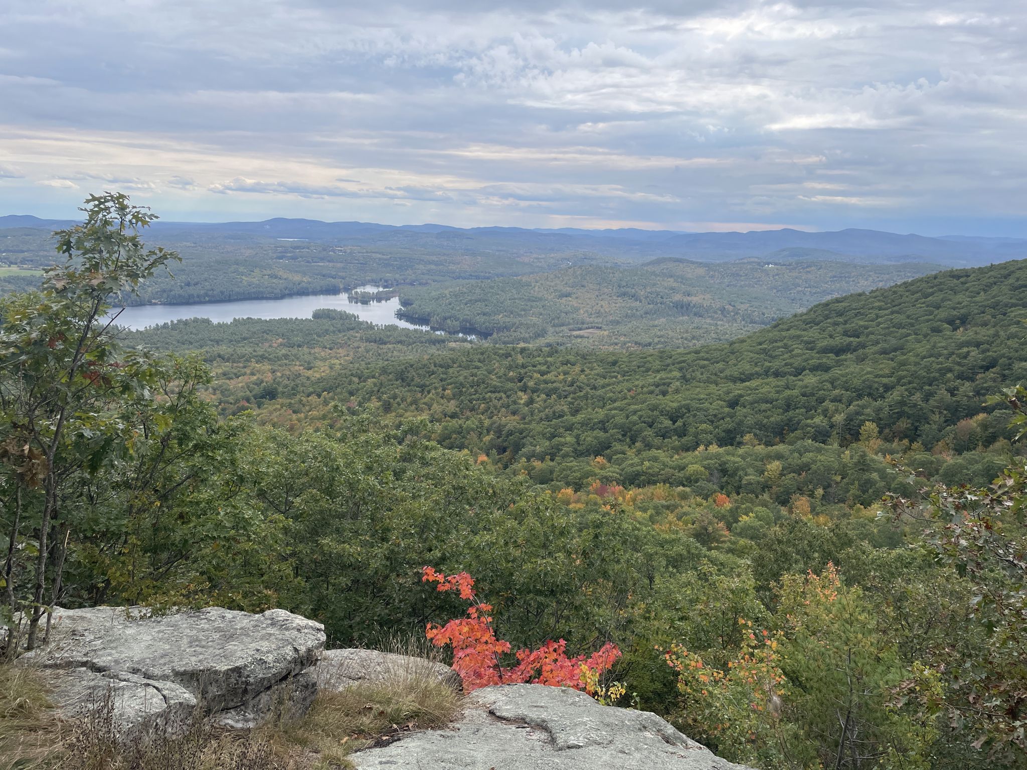 Rocky overlook with vibrant red foliage, green forest, and distant lake. - Ledges Trail