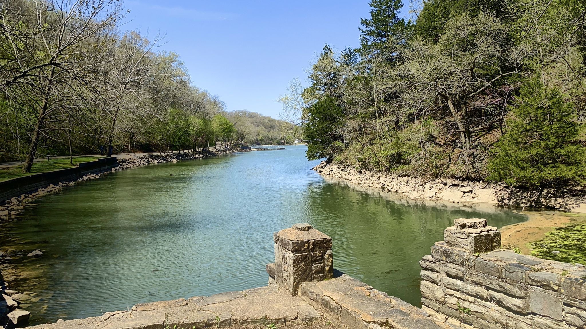 Calm river with tree-lined banks and stone structures under bright daylight. - Spring Trail