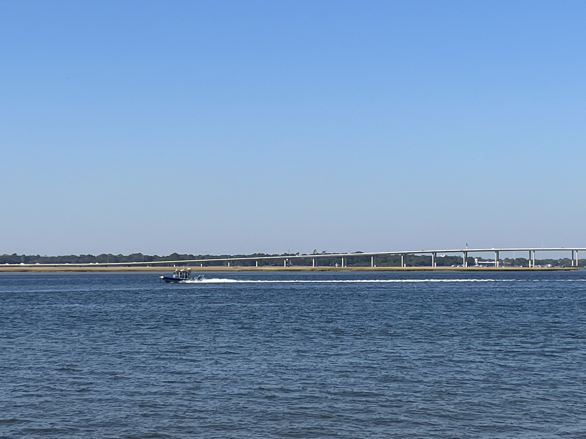 A boat moves across wide blue water with a long bridge in the distance. - Low Battery Trail
