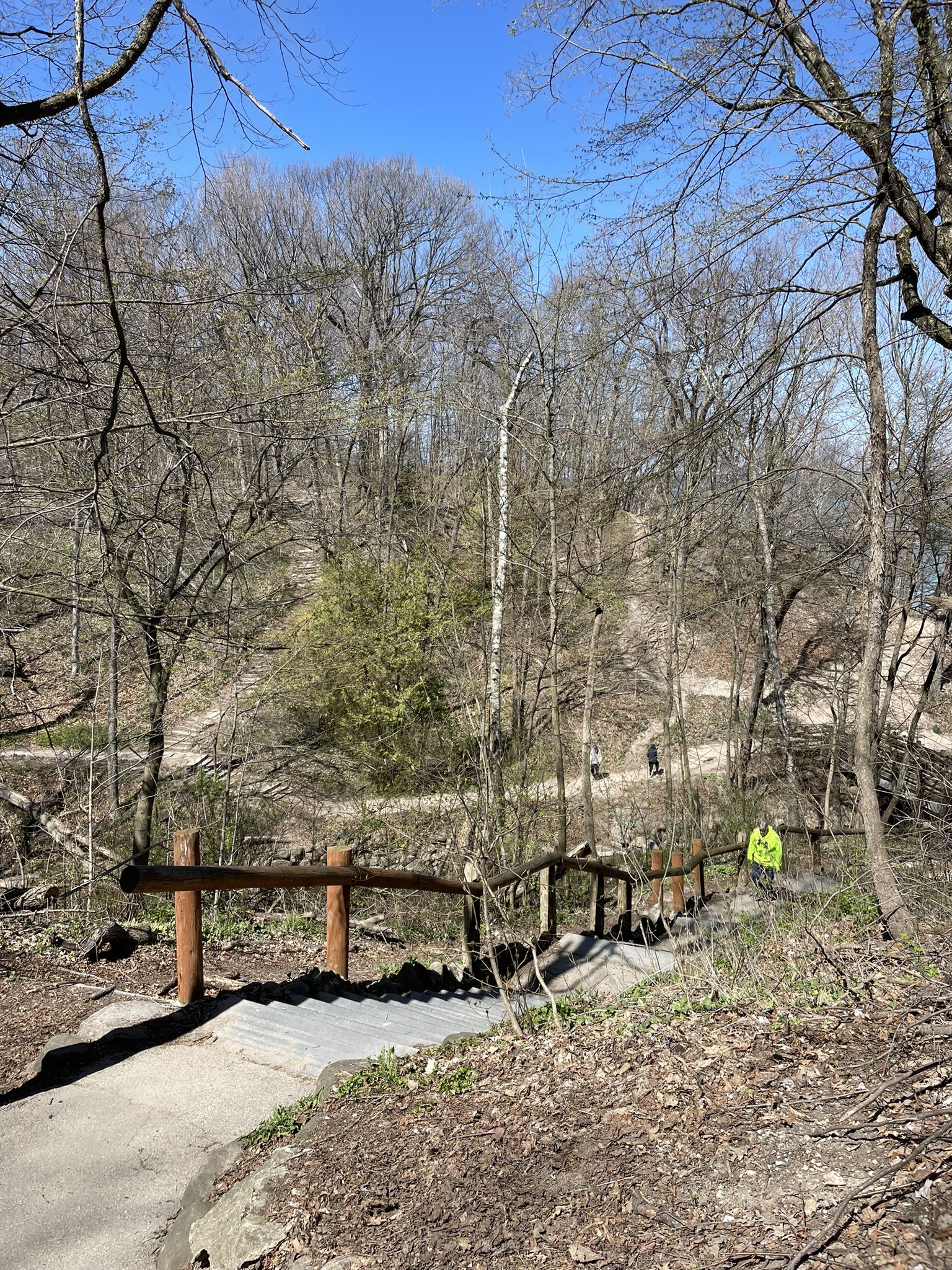 A person in a yellow jacket walks down a sunny, wooded path with wooden railings. - Seven Bridges Trail