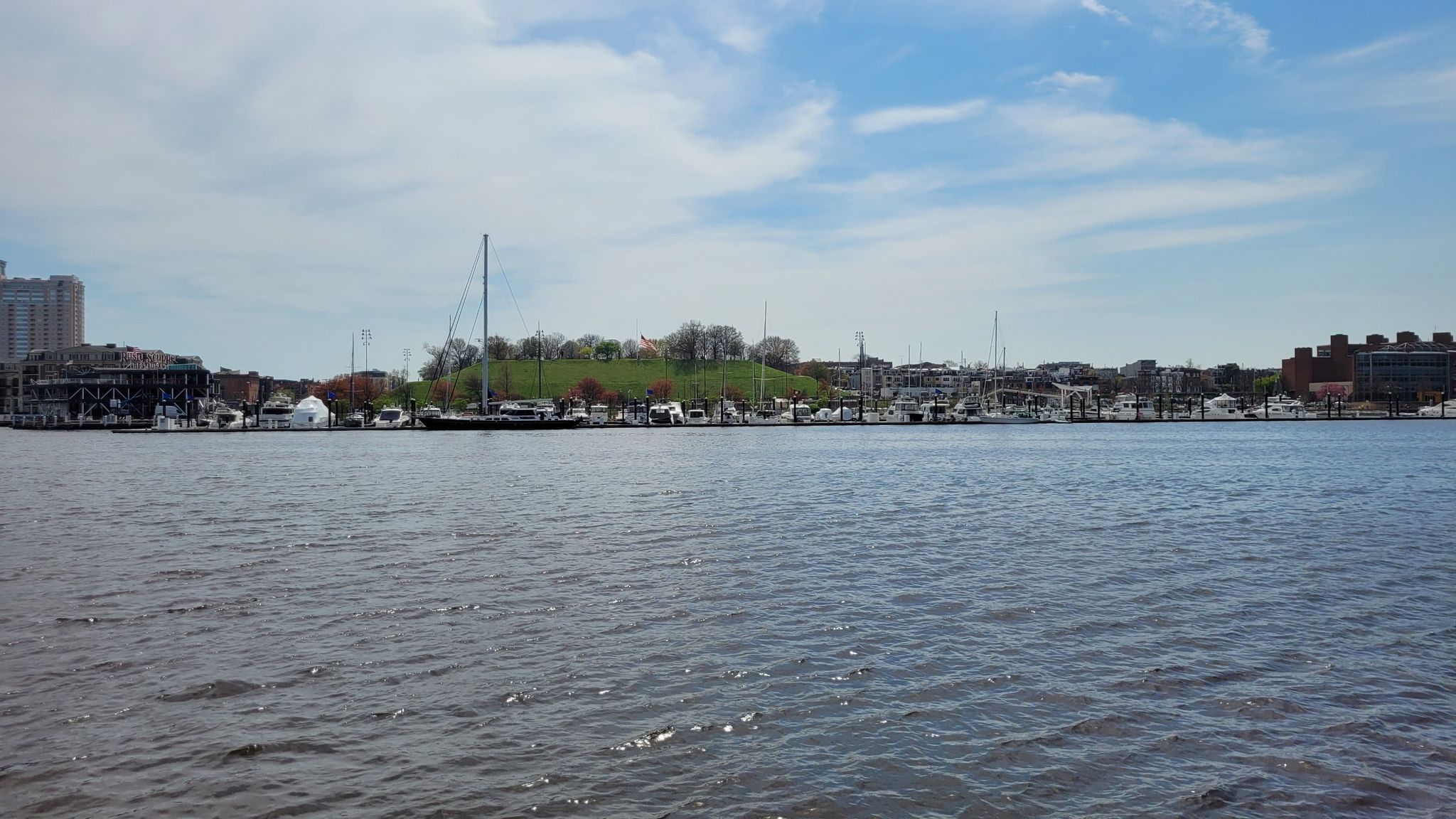 Wide view of a calm harbor with boats and a distant city skyline under clouds. - Baltimore Waterfront Promenade