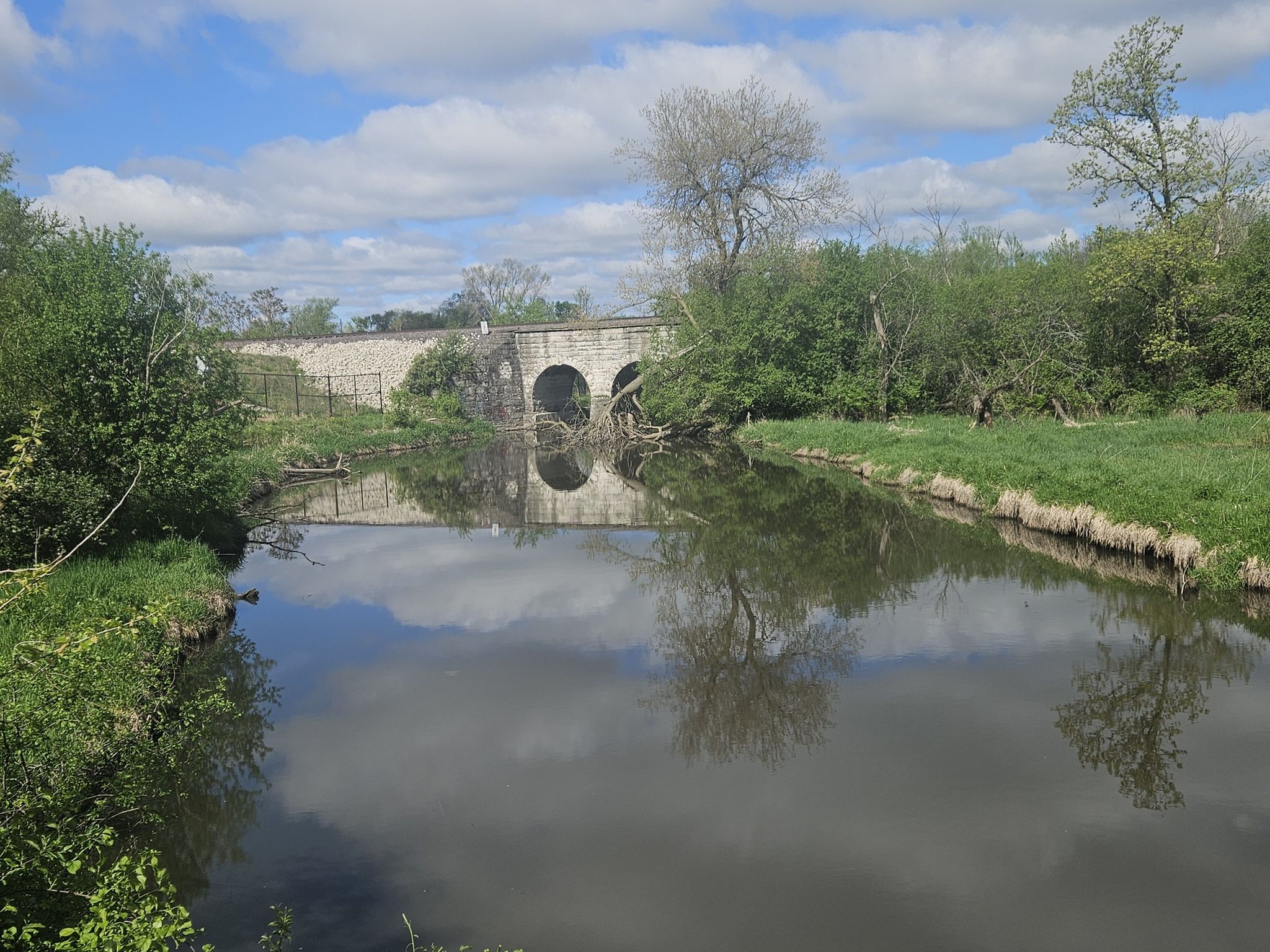 A stone arch bridge spans a calm river with green banks under a cloudy sky. - Norton Woods – Fox River Trail loop from St. Charles