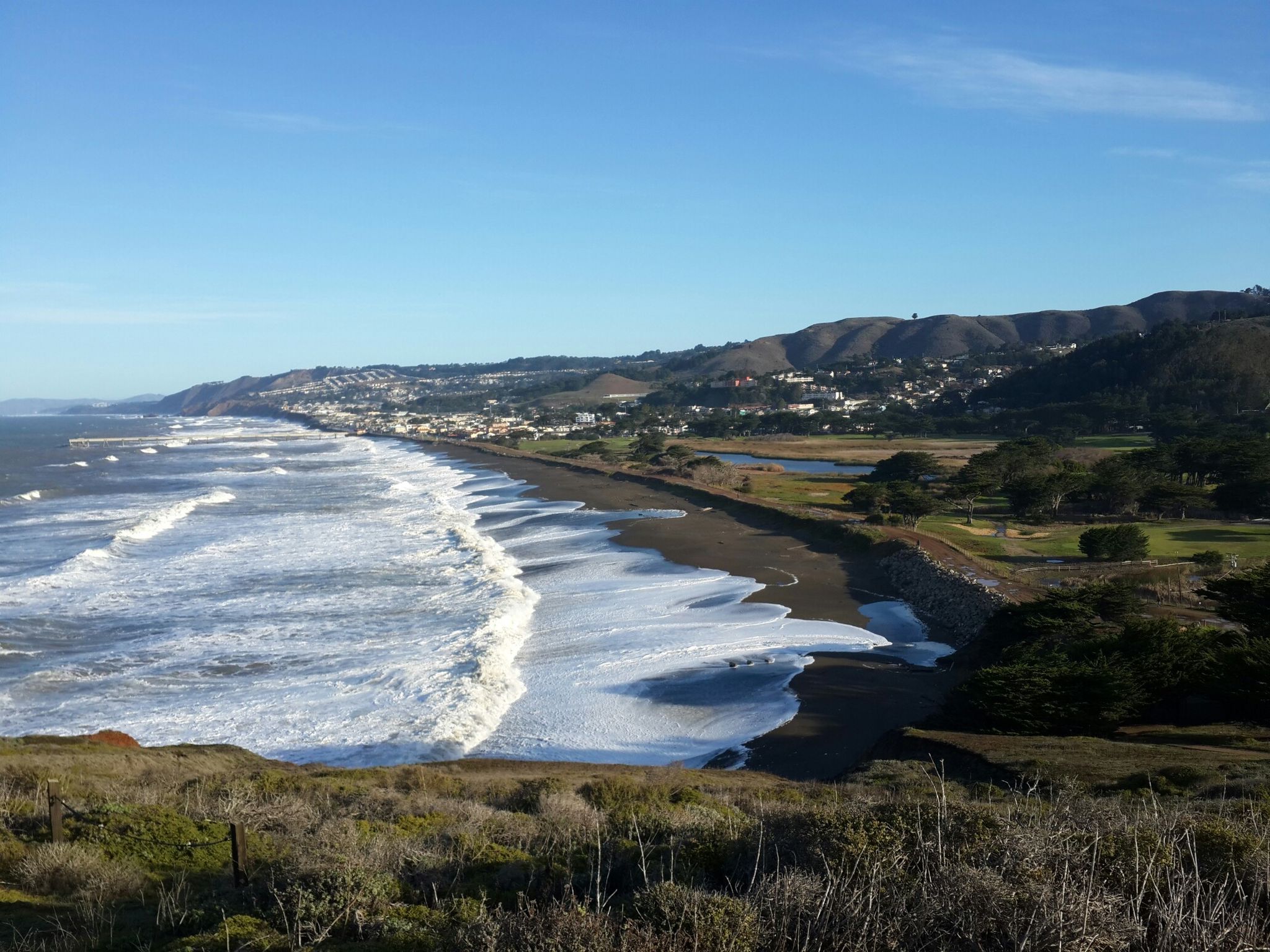 Waves crash on a long sandy beach next to a coastal town and green hills. - Mori Point Headlands – The Point loop from Milagra Ridge